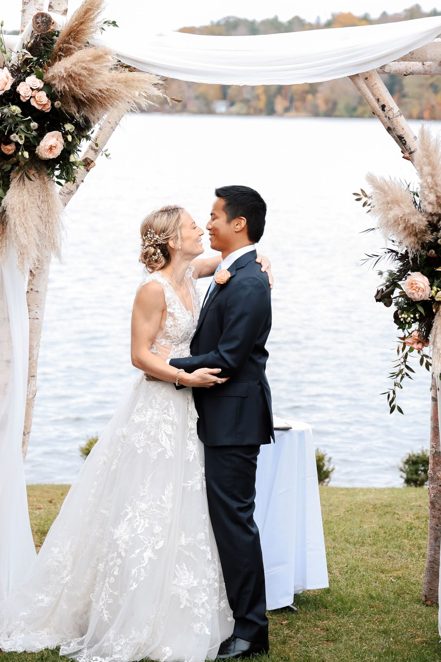 A romantic outdoor wedding ceremony by a lake with floral arrangements and white drapery decorations.