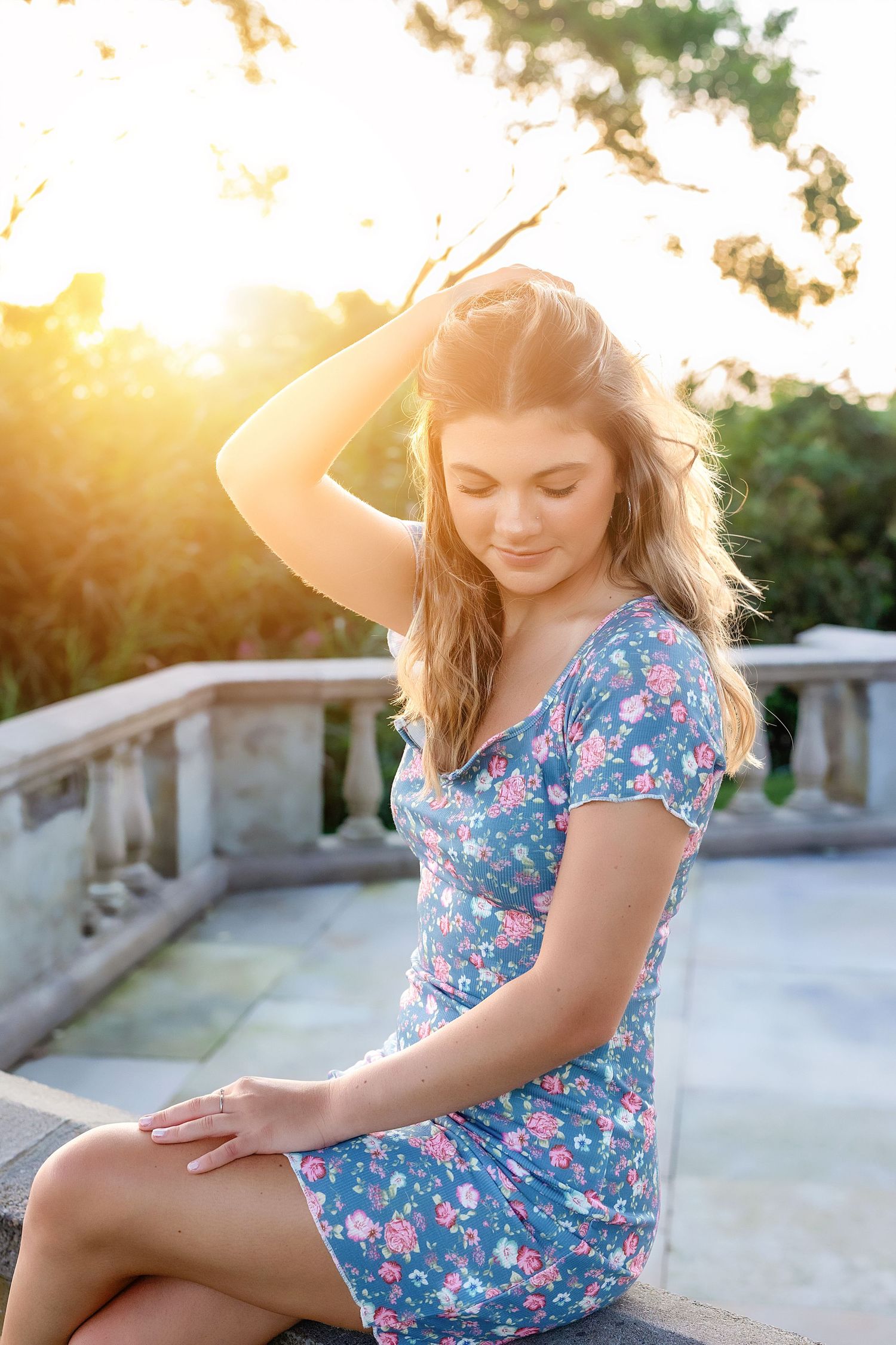A woman in a floral dress sits on a stone wall during golden hour sunset lighting.
