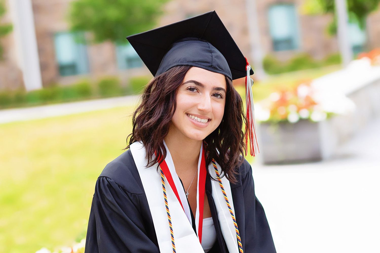 Graduate in traditional black cap and gown with honor cords stands smiling outdoors on college campus.