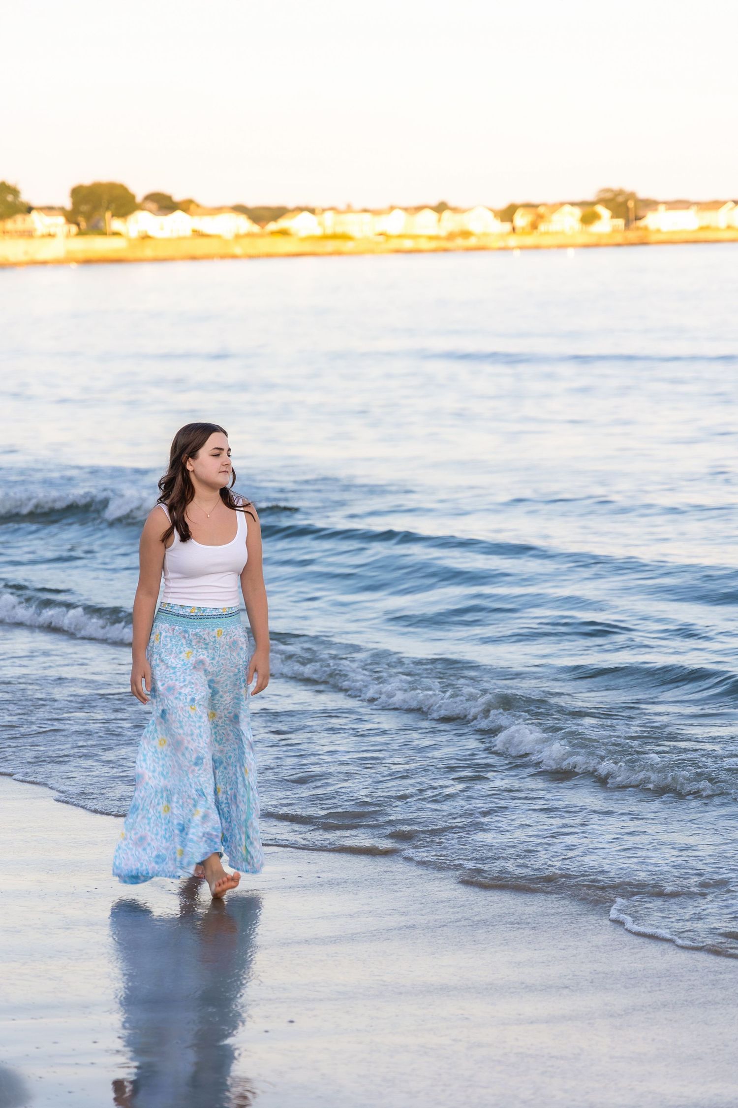 A person in a white top and floral blue skirt twirls playfully along the ocean shoreline at sunset.