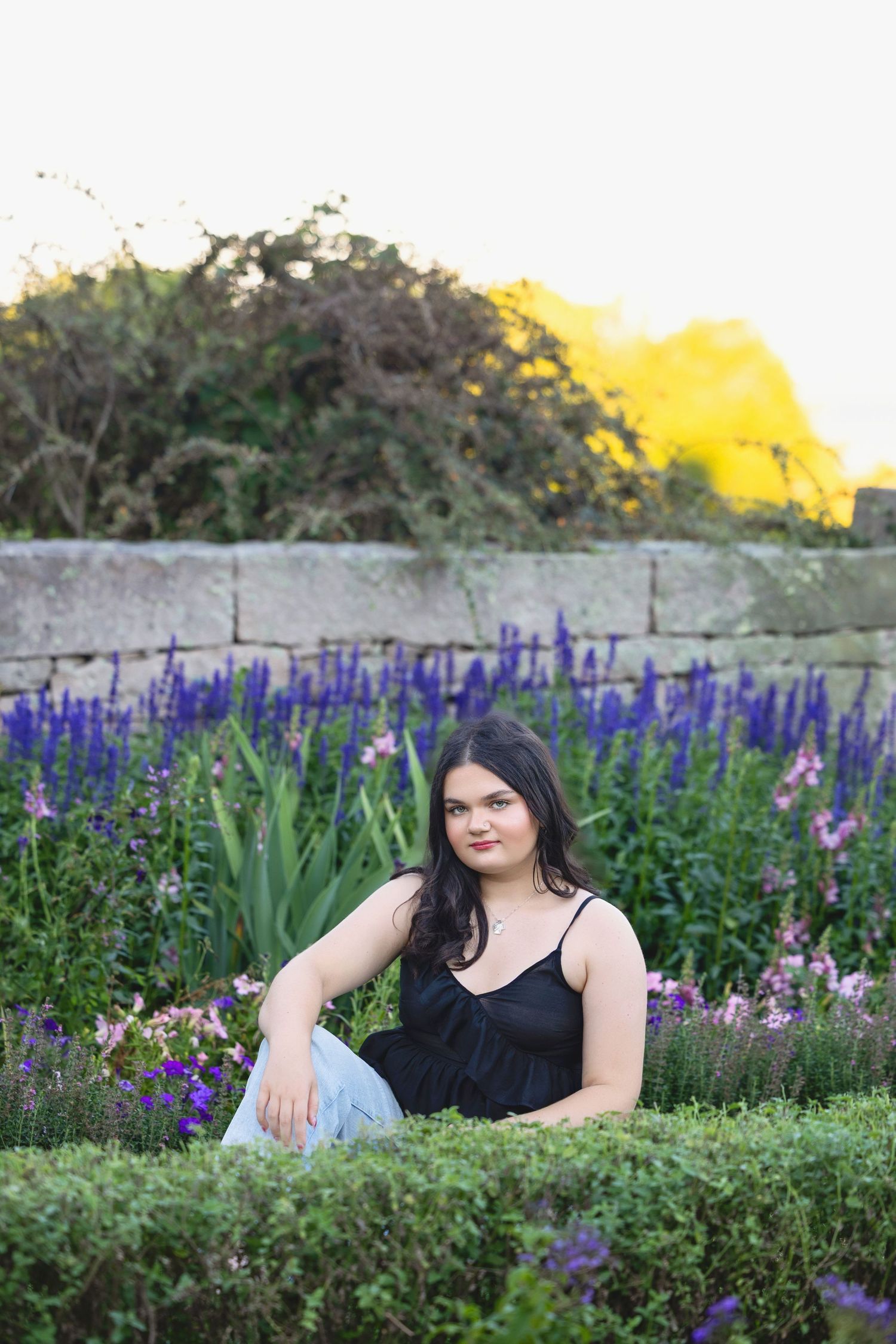 Someone sitting in a garden with blooming purple delphiniums and a stone wall backdrop.