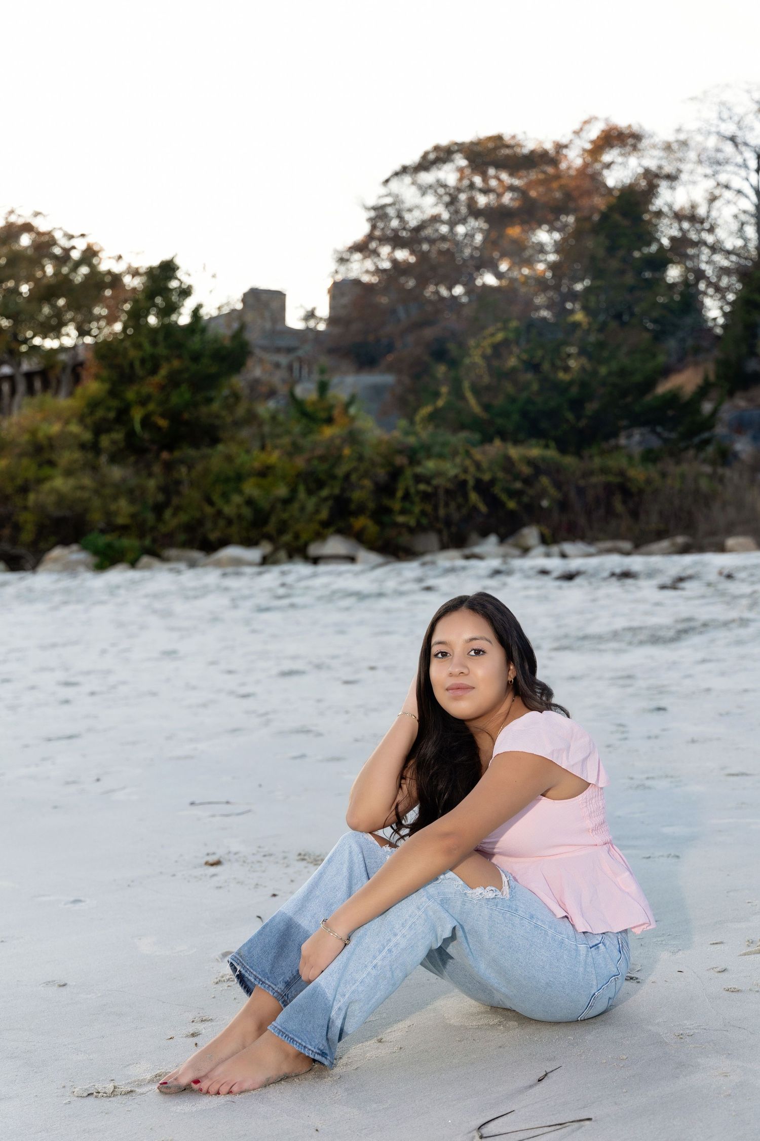 A person in jeans and pink top lounges on a sandy beach during sunset, creating a serene coastal scene.