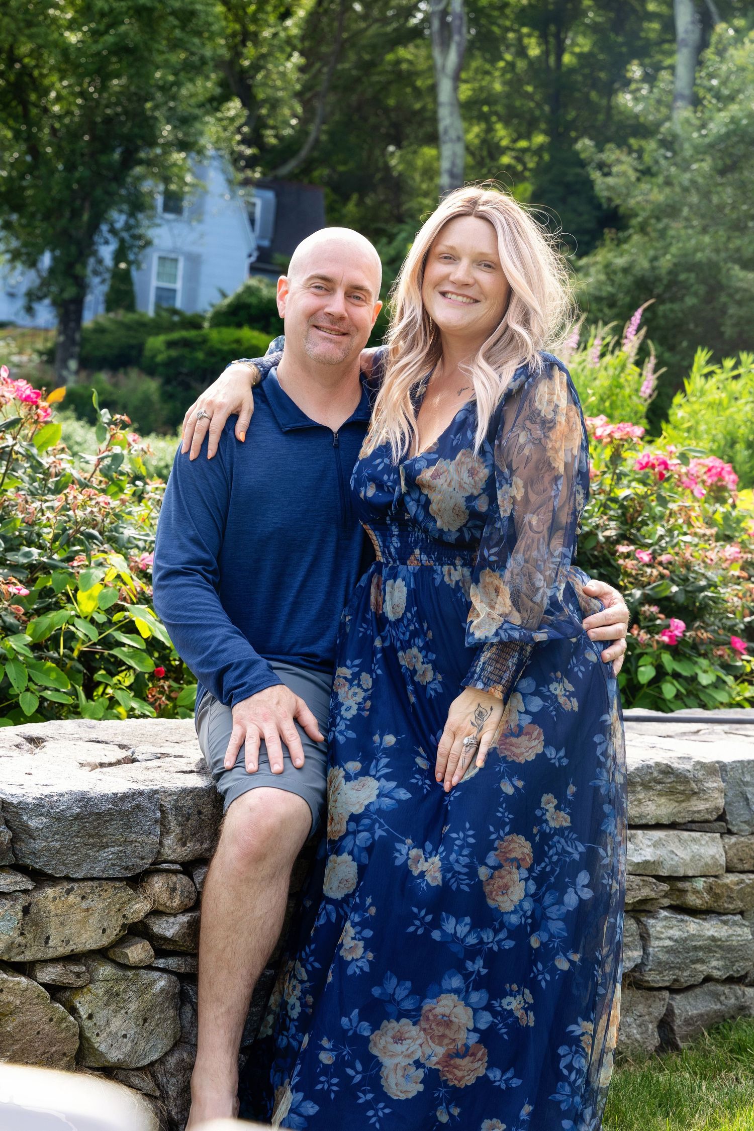 A couple sits together on a stone wall in a garden with blooming flowers and greenery in the background.