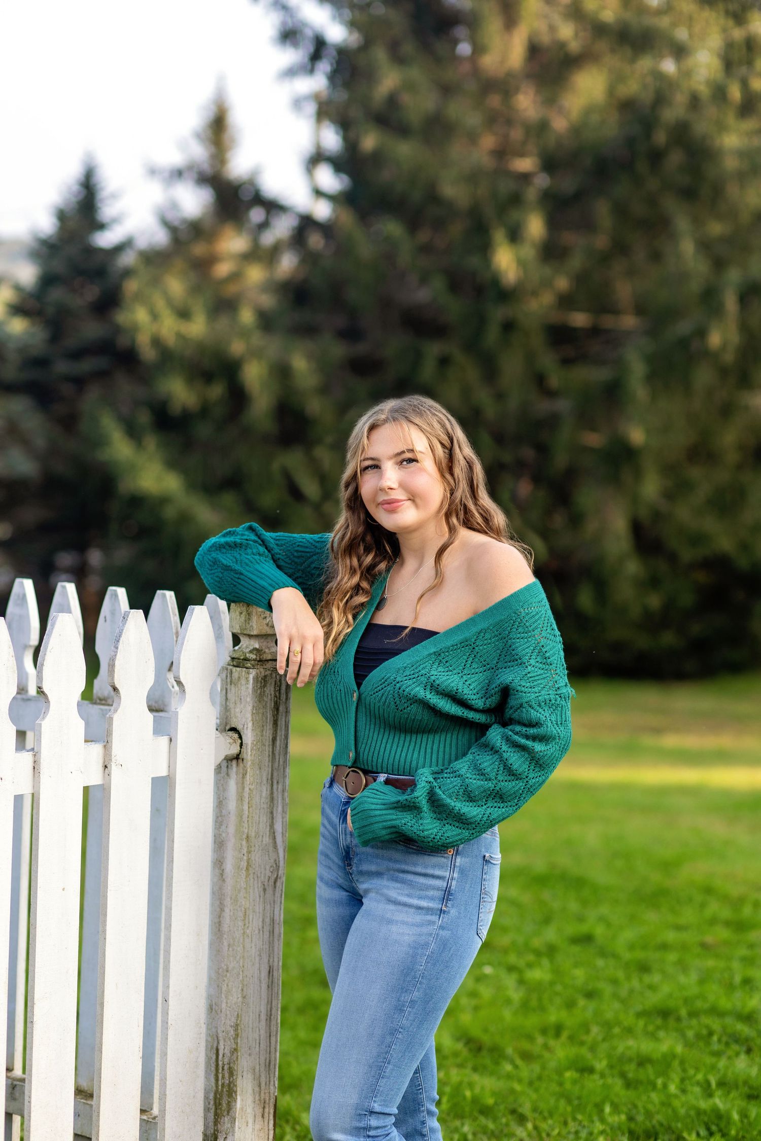 Woman in green sweater and jeans leans against white picket fence with evergreen trees in background.