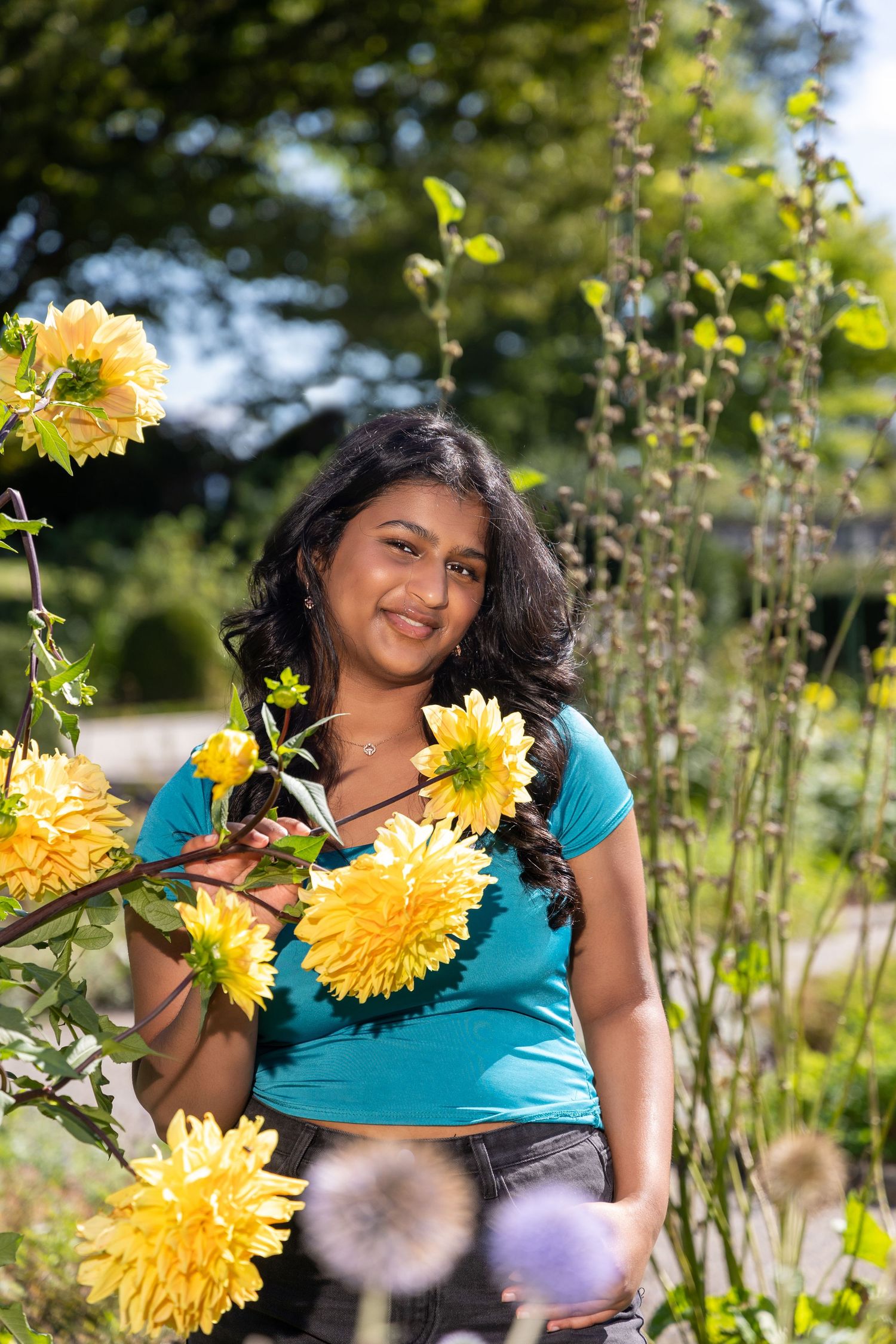 A woman in a turquoise top poses joyfully among blooming yellow sunflowers in a garden setting.