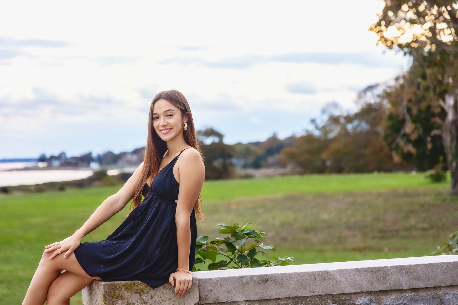 Woman in black dress sits gracefully on wooden bench in scenic outdoor garden setting with natural lighting.