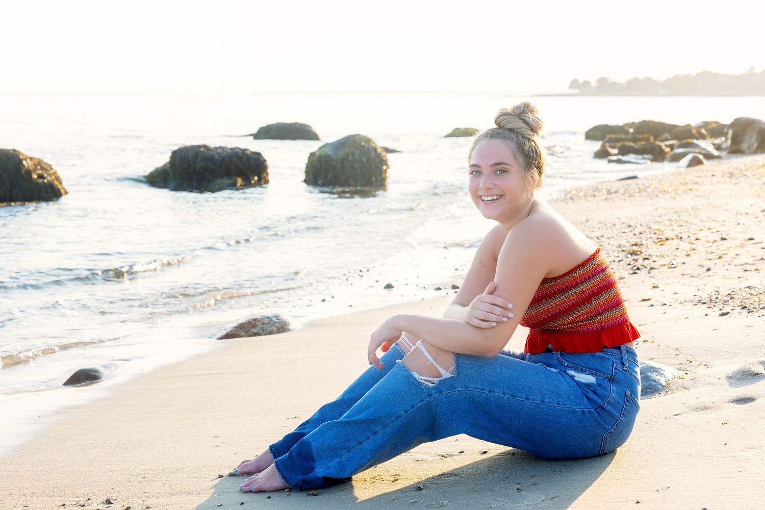 Young woman in red tank top and jeans sits on sandy beach with ocean waves and rocks in background.