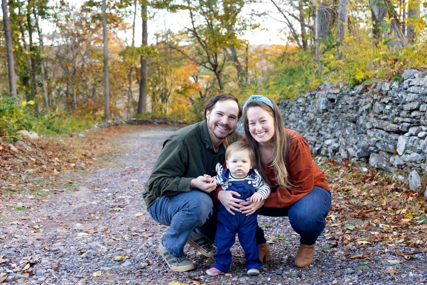 A family poses together on an autumn path lined with fallen leaves and a stone wall in the background.