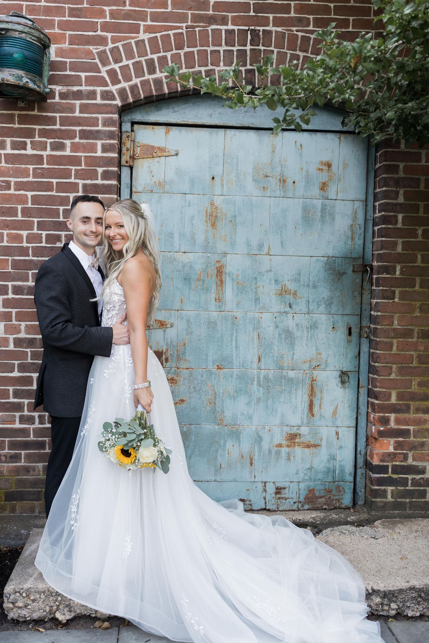 A wedding couple poses against a rustic blue door and brick wall with sunflowers in a romantic photo series.