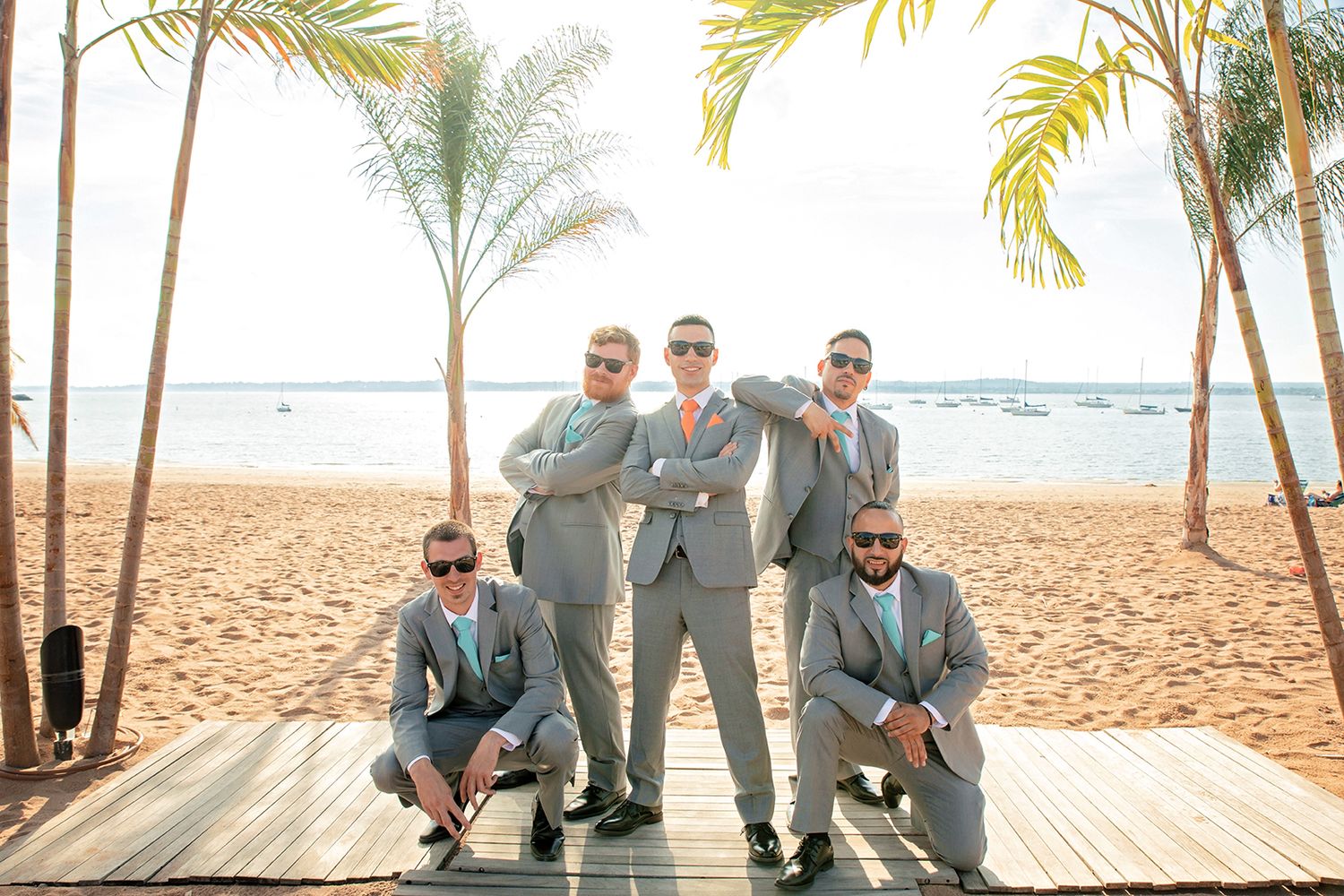 Wedding party in gray suits poses together on a tropical beach with palm trees and ocean views.