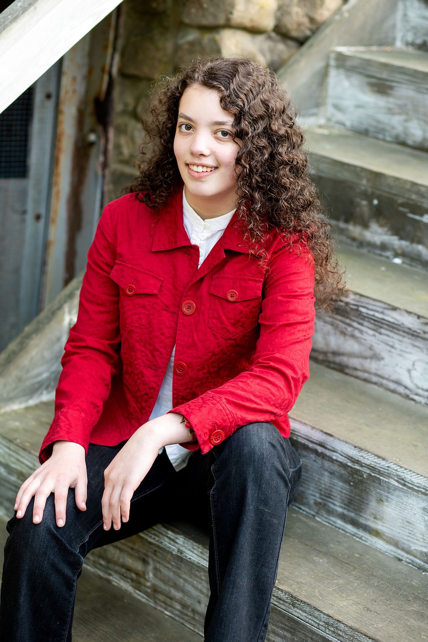 A person with long curly hair wearing a bright red jacket sits on stone steps outdoors.