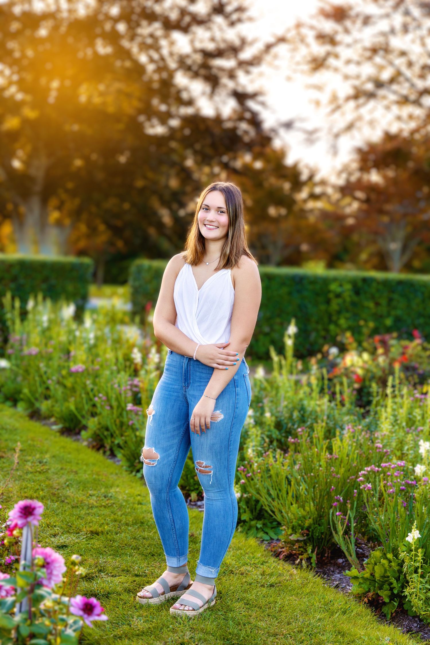 Someone stands smiling in a garden with blooming flowers and hedges during golden hour.