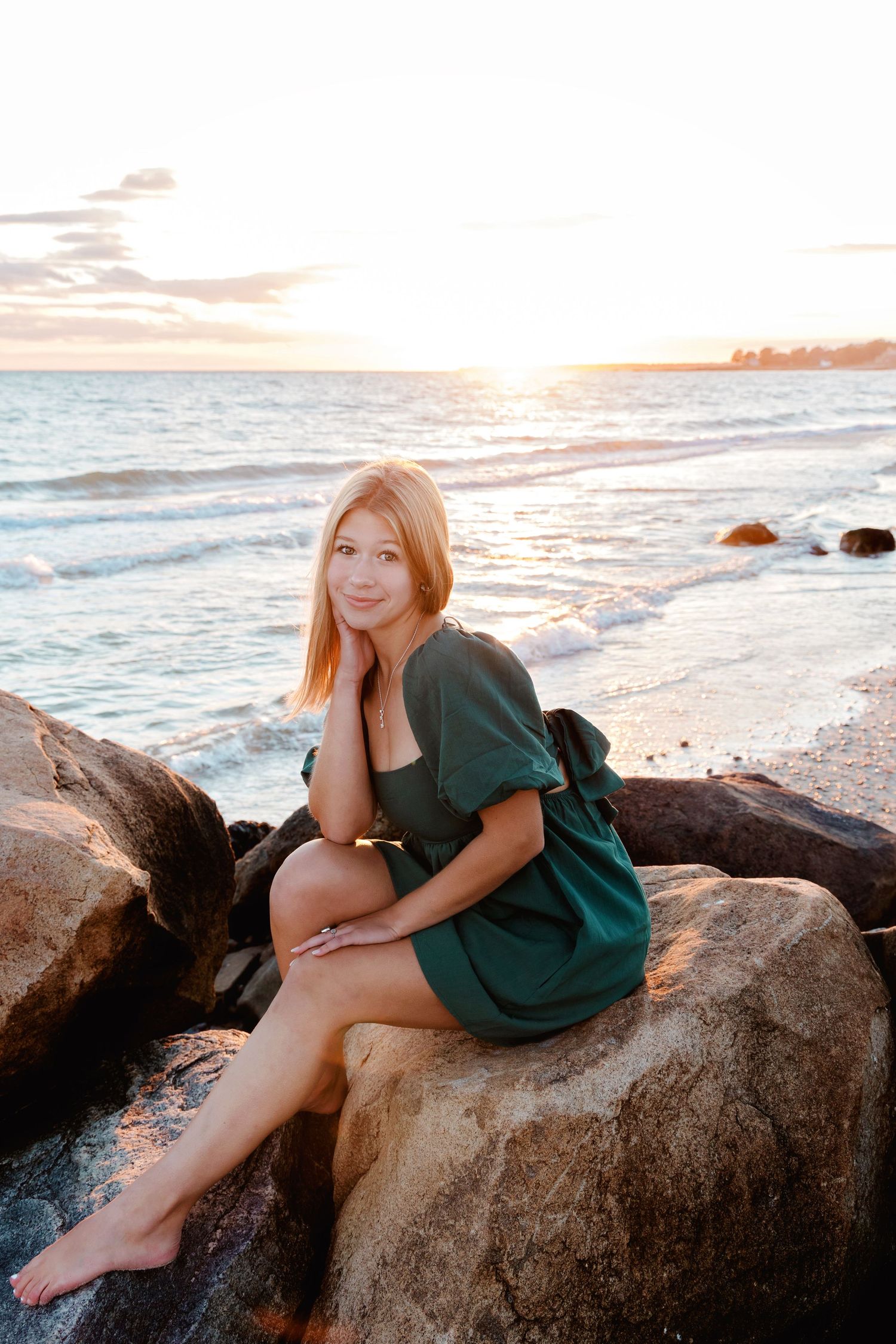 A person in a green dress reclines on coastal rocks at sunset by the ocean.