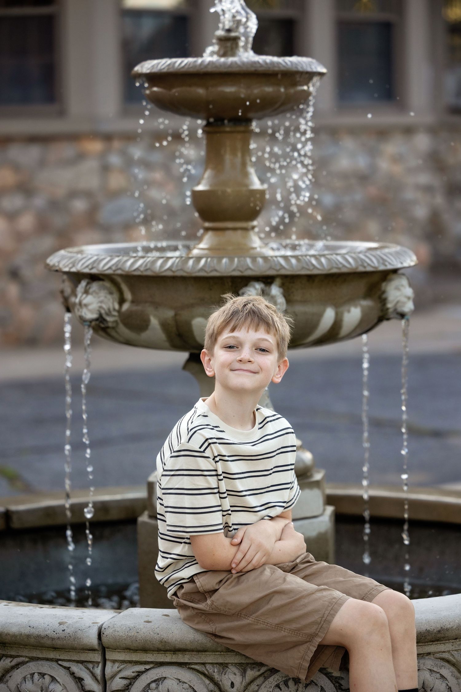 A young child sits playfully by a decorative stone fountain with cascading water in an outdoor setting.