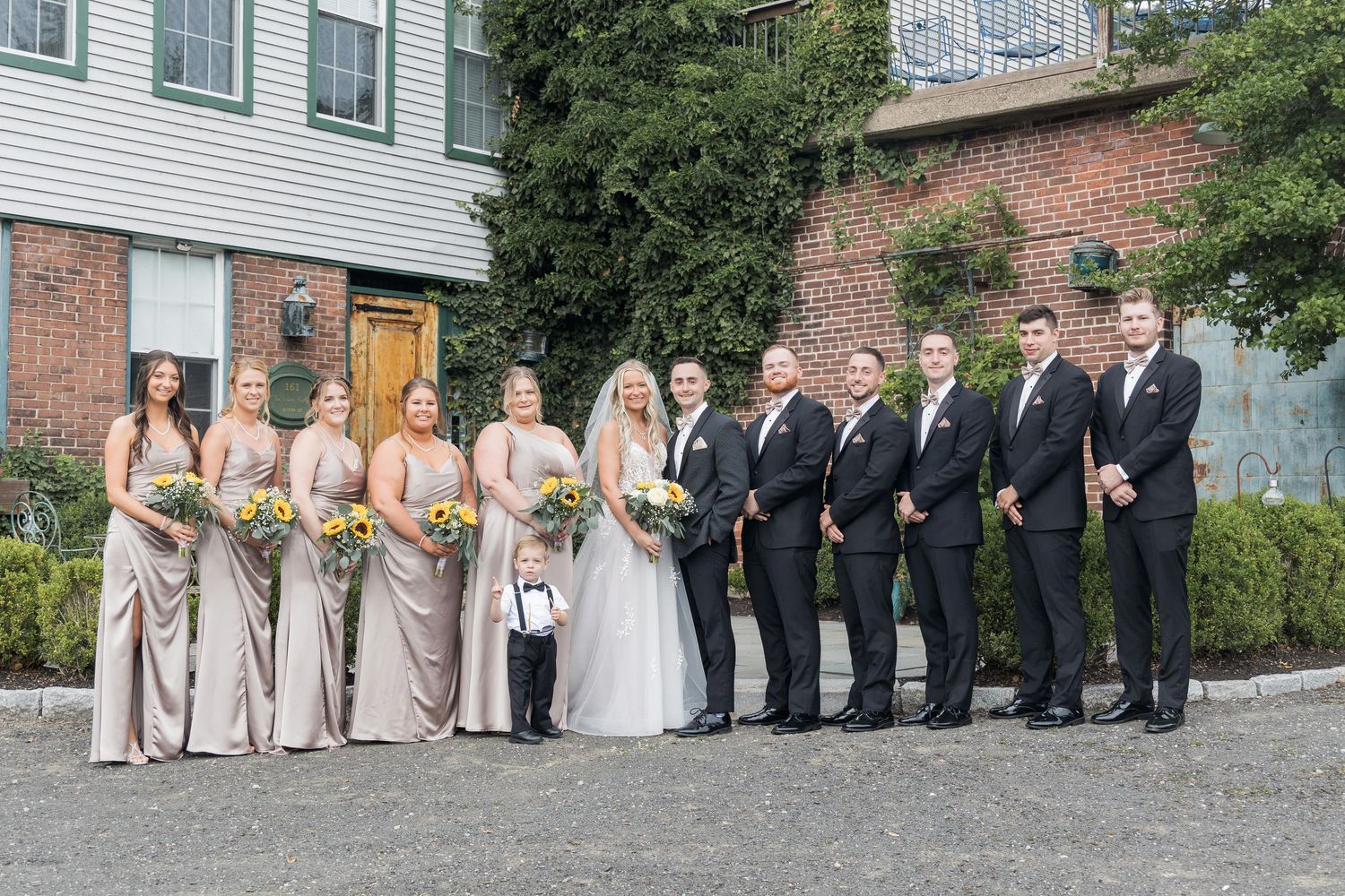 Large wedding party poses together on gravel path with bridesmaids in neutral dresses and groomsmen in black suits.