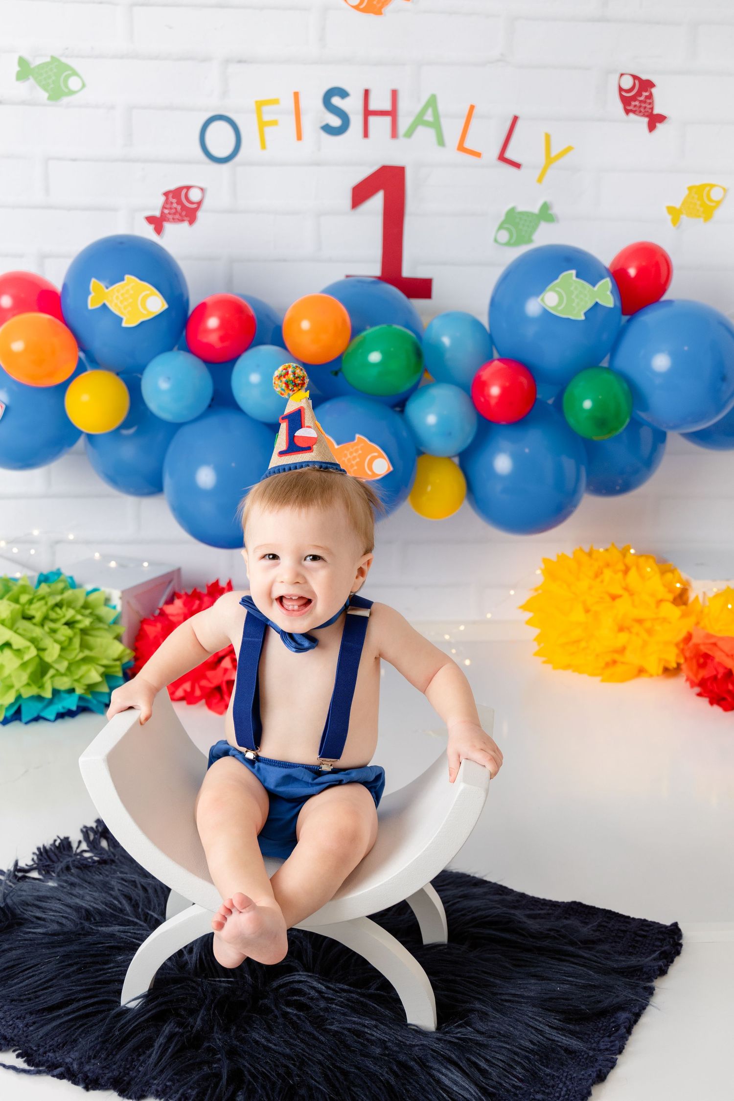 First birthday party decoration with colorful balloon arch, fish theme, and navy blue rug on white background.