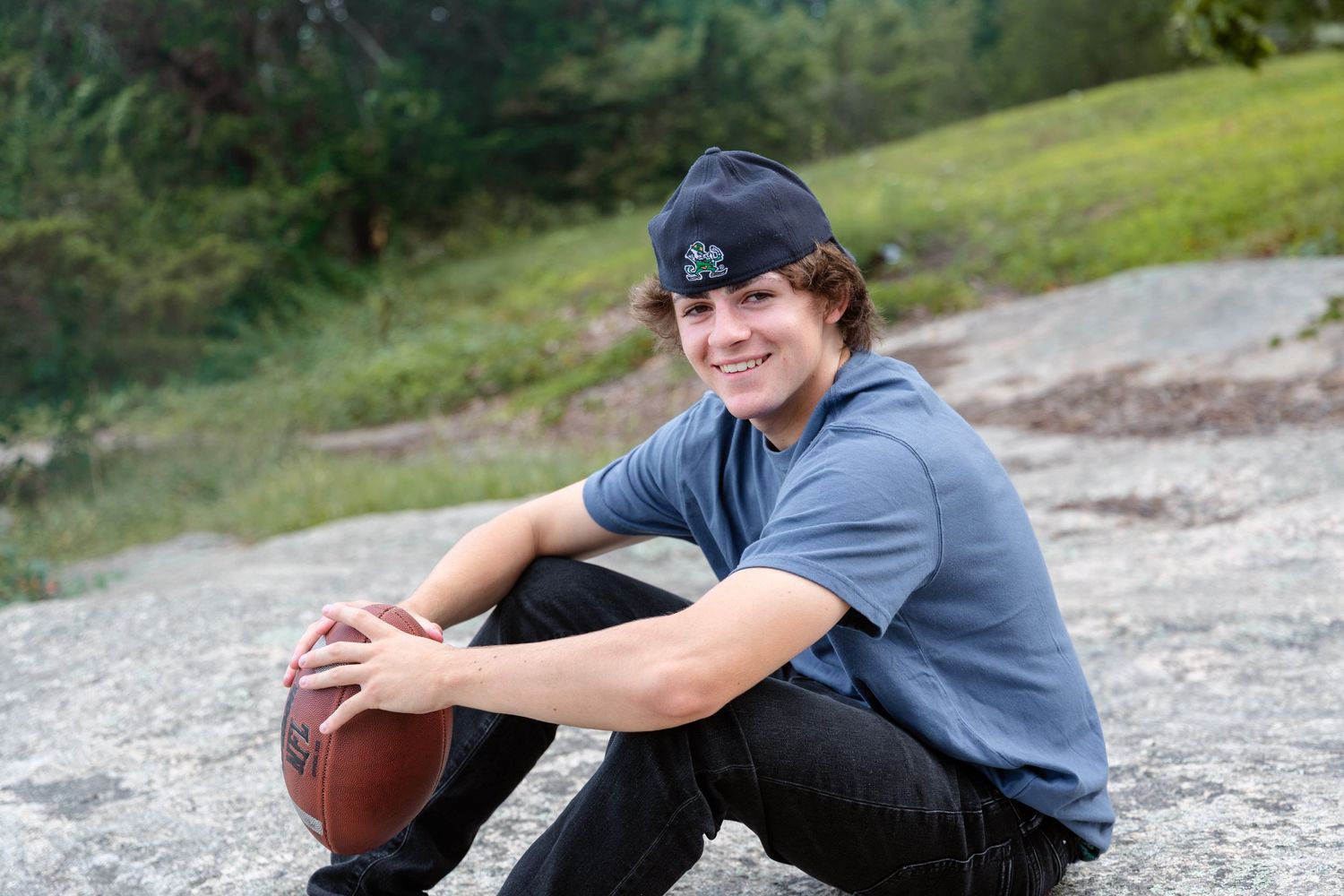 A person in a gray shirt sits outdoors holding a football on a gravel path with a grassy hill in the background.