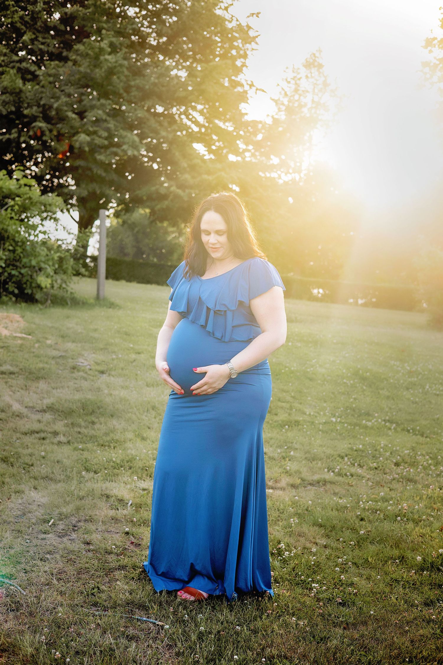 Maternity photoshoot in a grassy field at sunset with golden sunlight filtering through the trees.