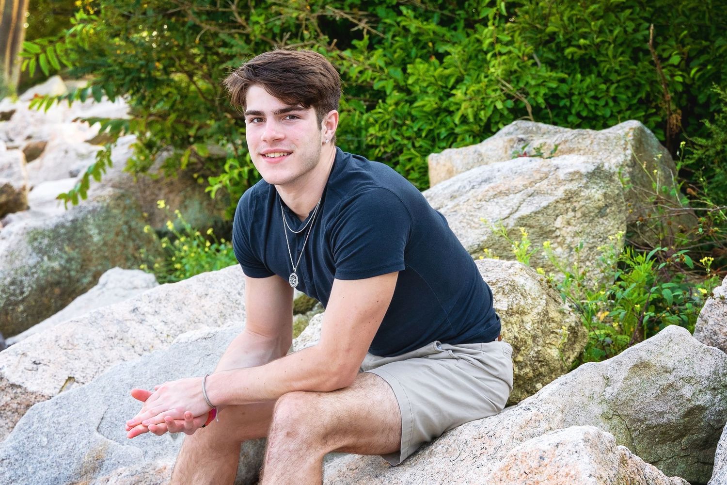 Someone in a black t-shirt and khaki shorts sits casually on large rocks in a natural outdoor setting.