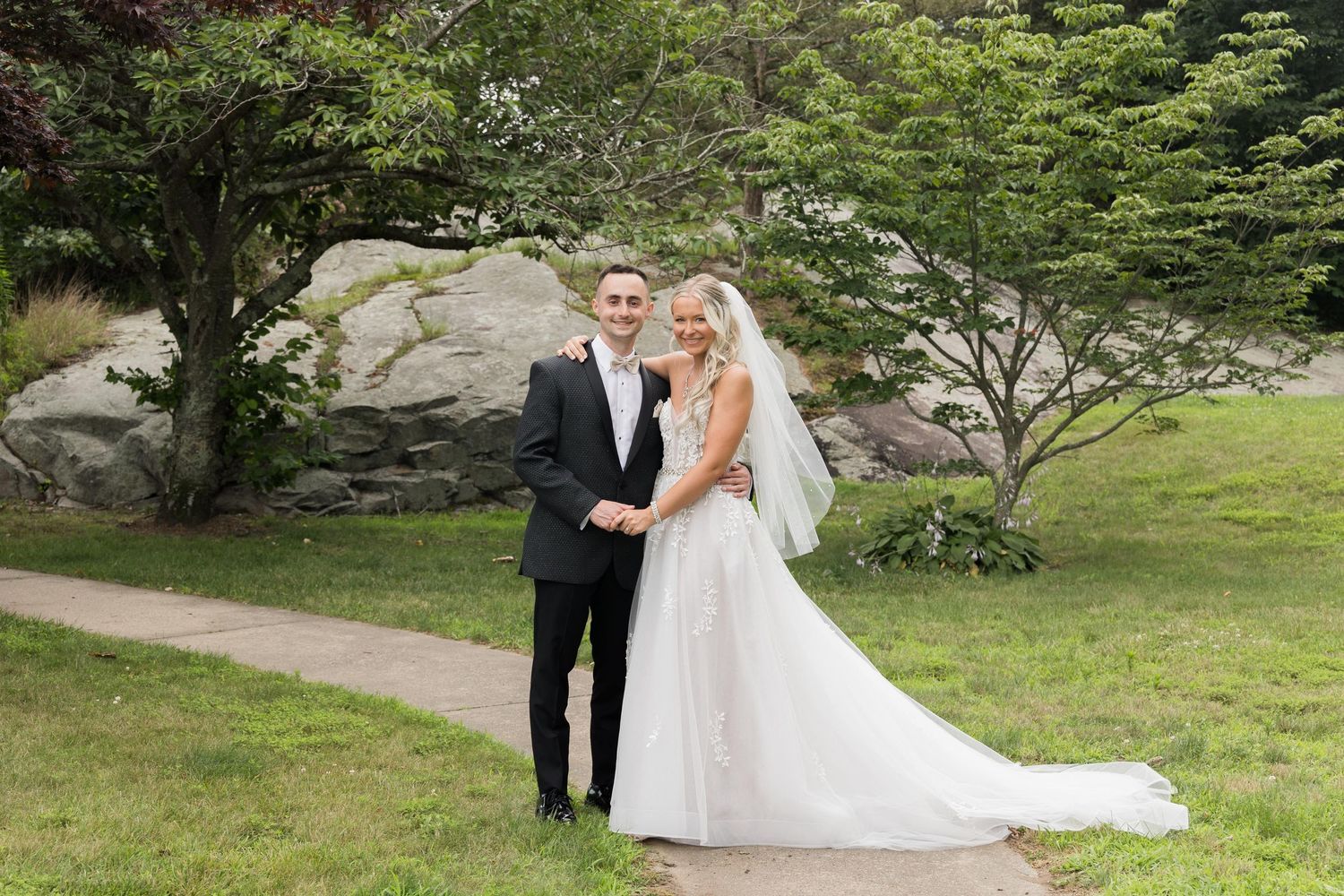 A wedding couple poses in a lush garden setting with a flowing white gown and classic black tuxedo.