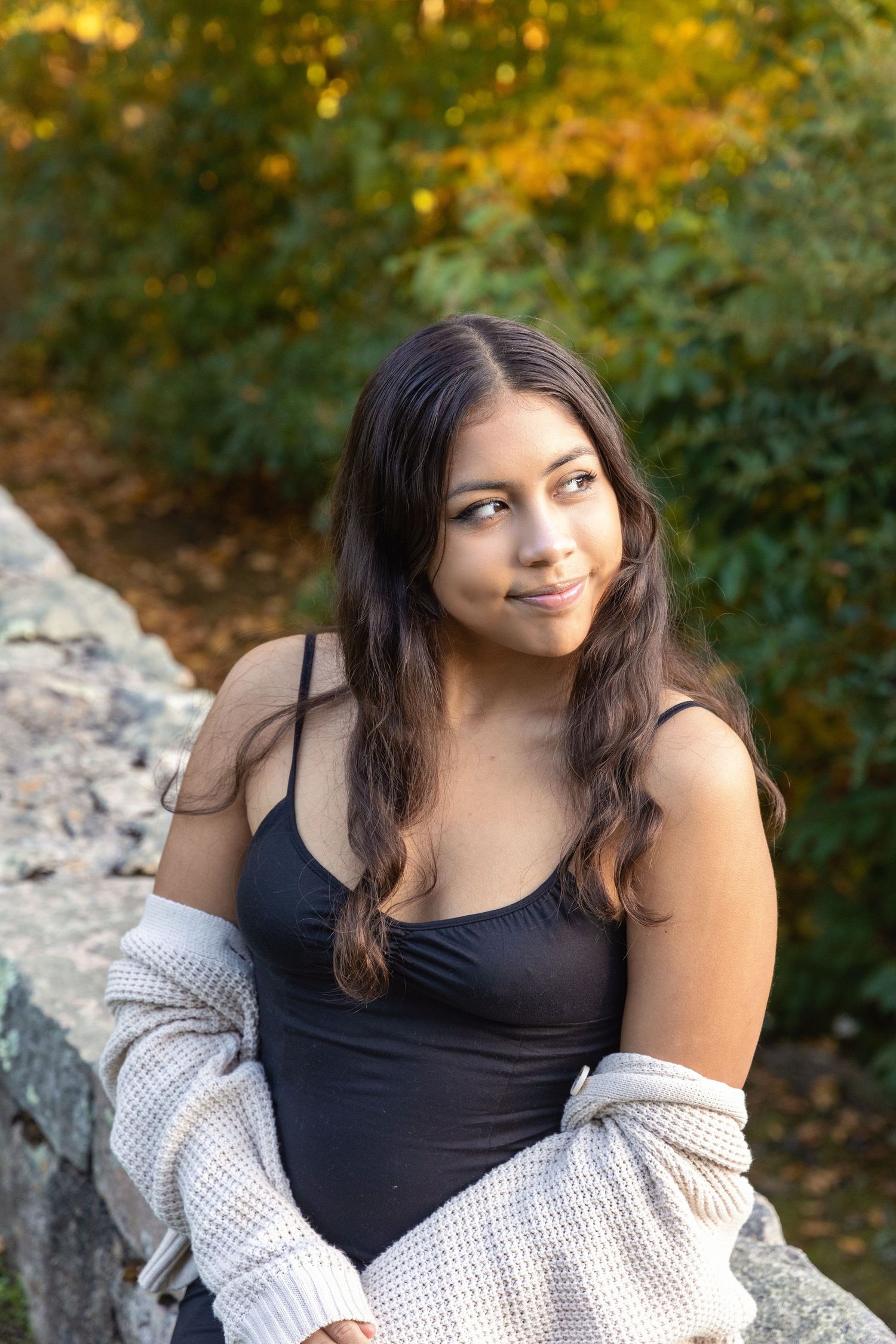 Portrait of someone in a black tank top and knit cardigan sitting on rocks with autumn foliage in the background.