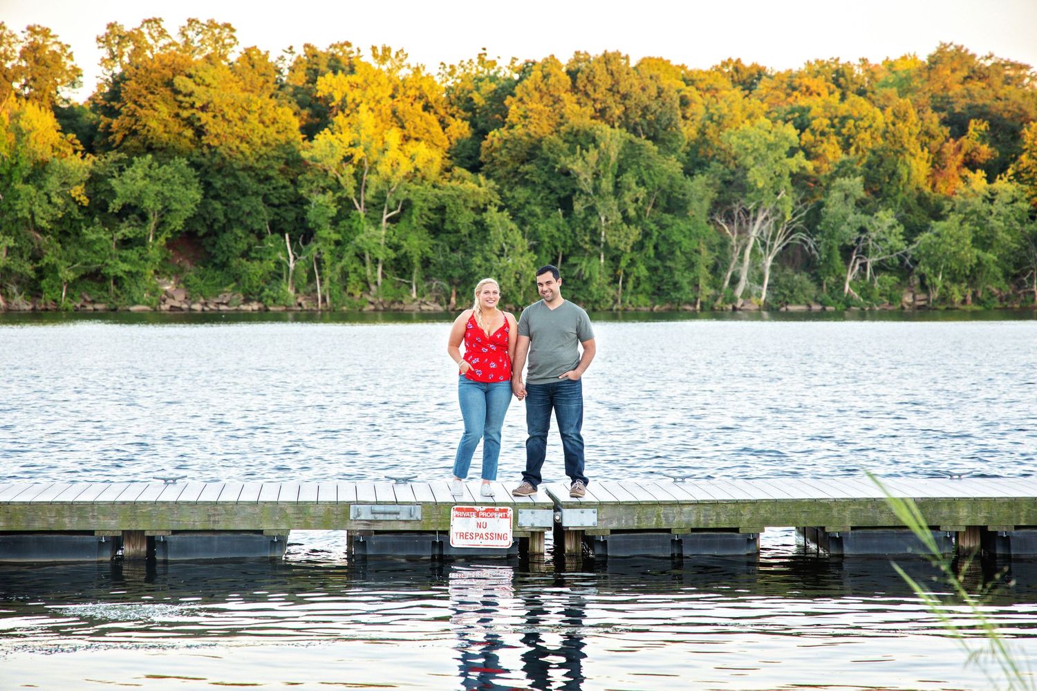 Two people stand at the edge of a lake surrounded by autumn trees with golden foliage reflecting in the water.