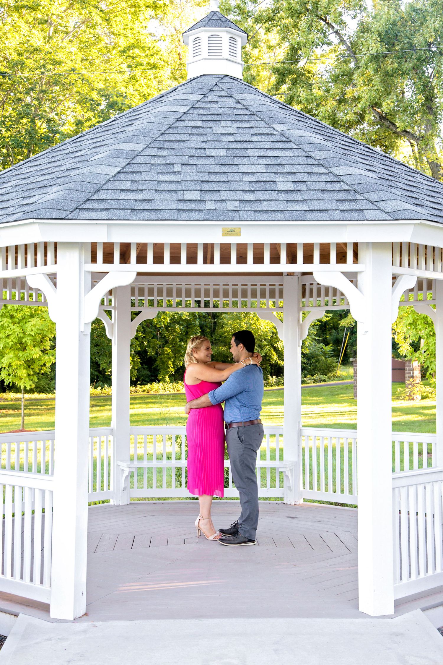 A couple dances together in a white wooden gazebo with a gray shingled roof surrounded by green trees in a park.