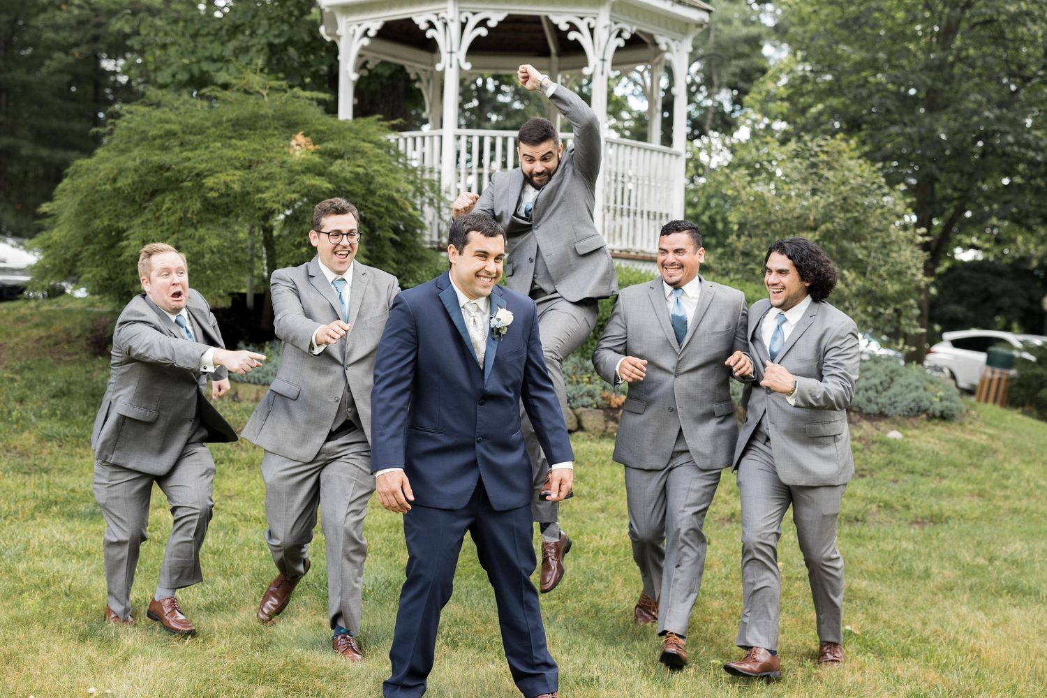 Wedding party members in gray and navy suits pose playfully for photos on a grassy hill with a gazebo in the background.