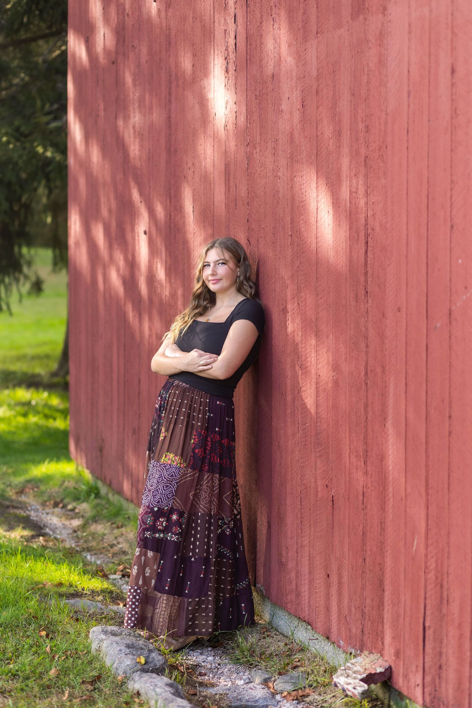 A figure in a black top and patterned maxi skirt leans against a rustic red barn wall in dappled sunlight.