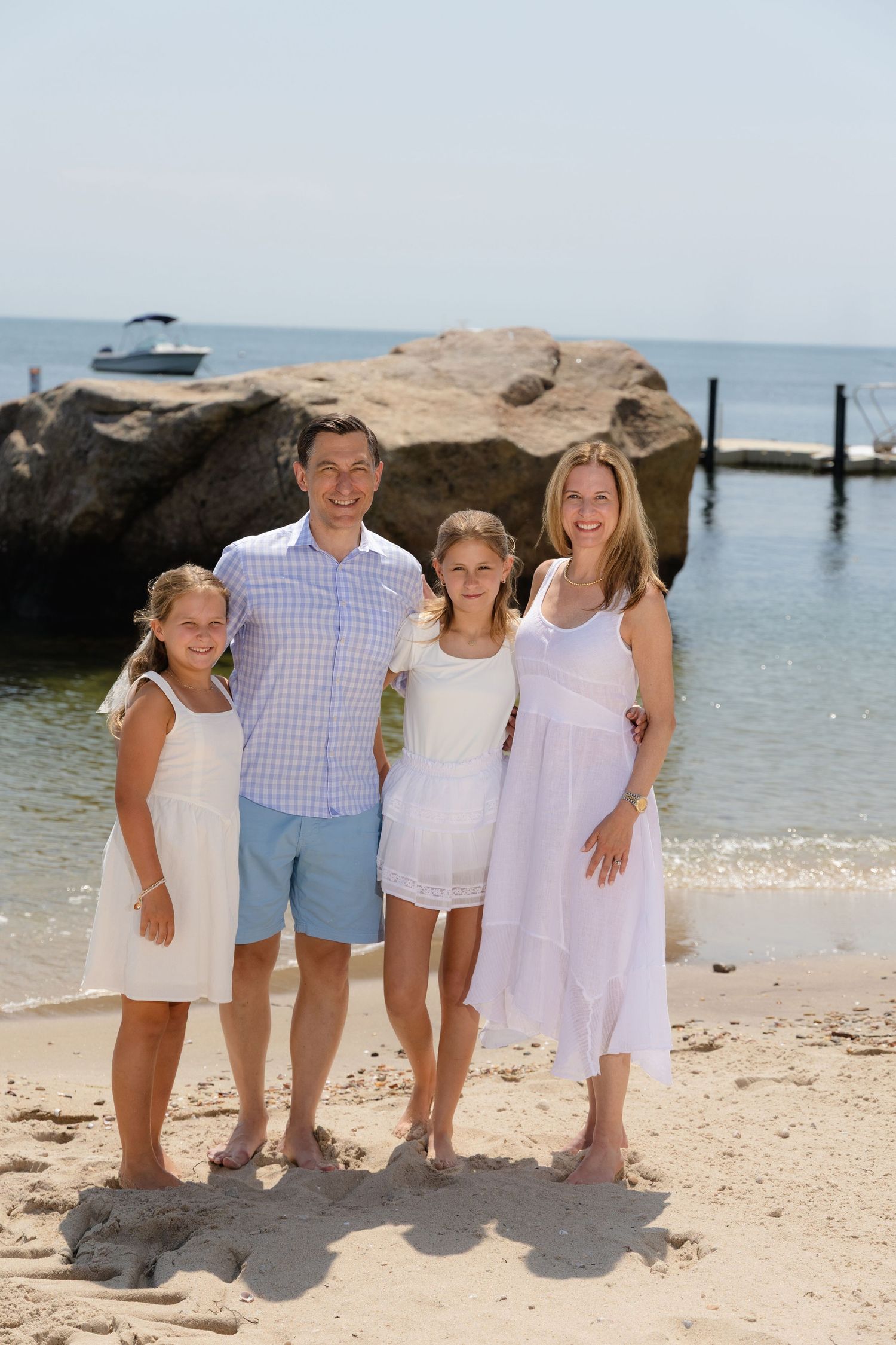 A family poses together on a sunny beach with boats and rocks visible in the background.