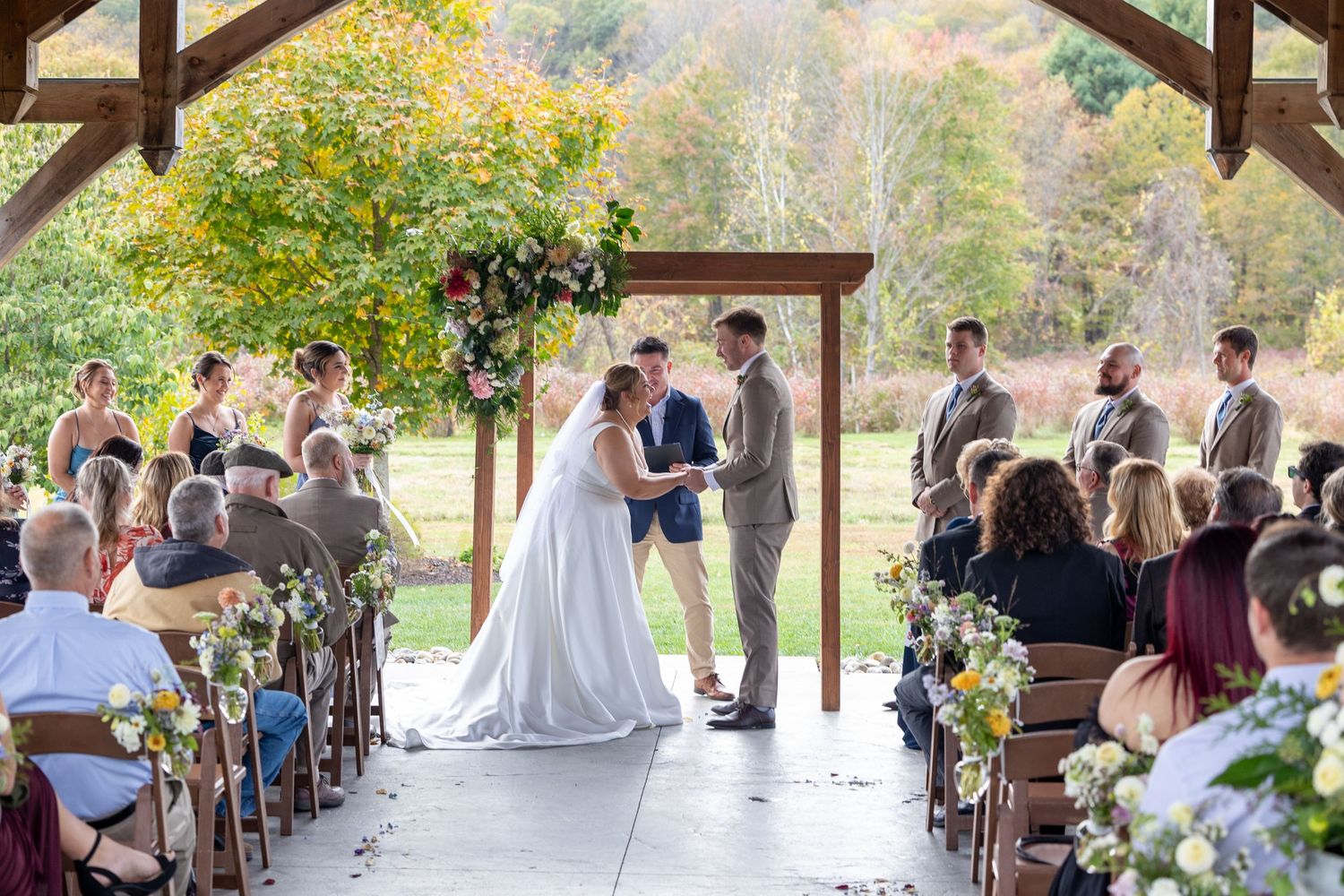 Outdoor covered pavilion wedding ceremony with fall foliage in background.