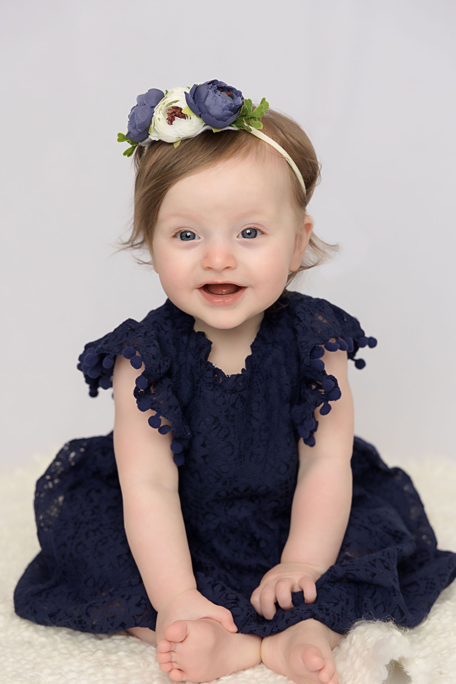 A baby wearing a navy blue lace dress sits on a white background with a floral headband.