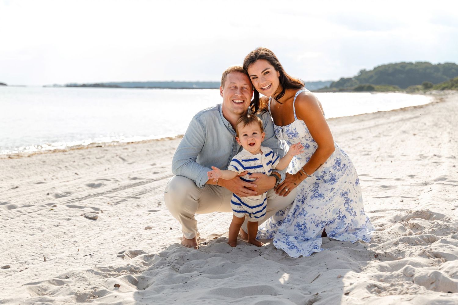 A family poses together on a sunny beach with ocean and coastline in the background.
