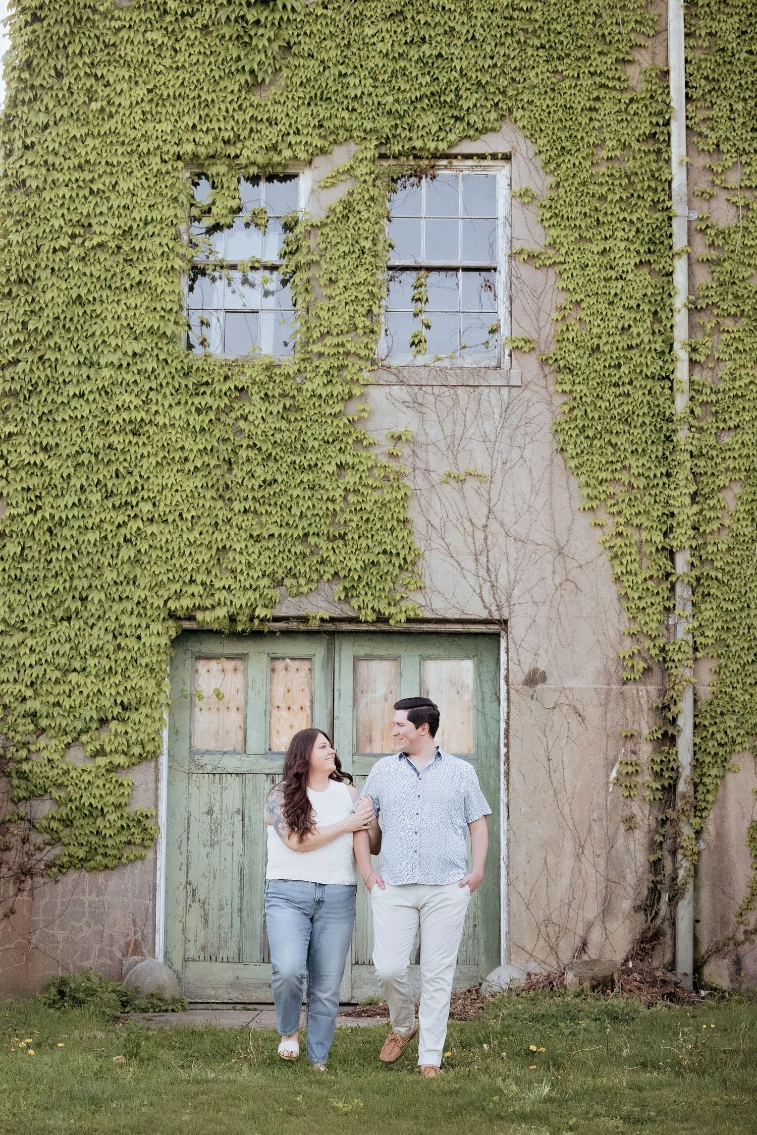 A couple stands together in front of an ivy-covered wall with vintage green doors and windows.