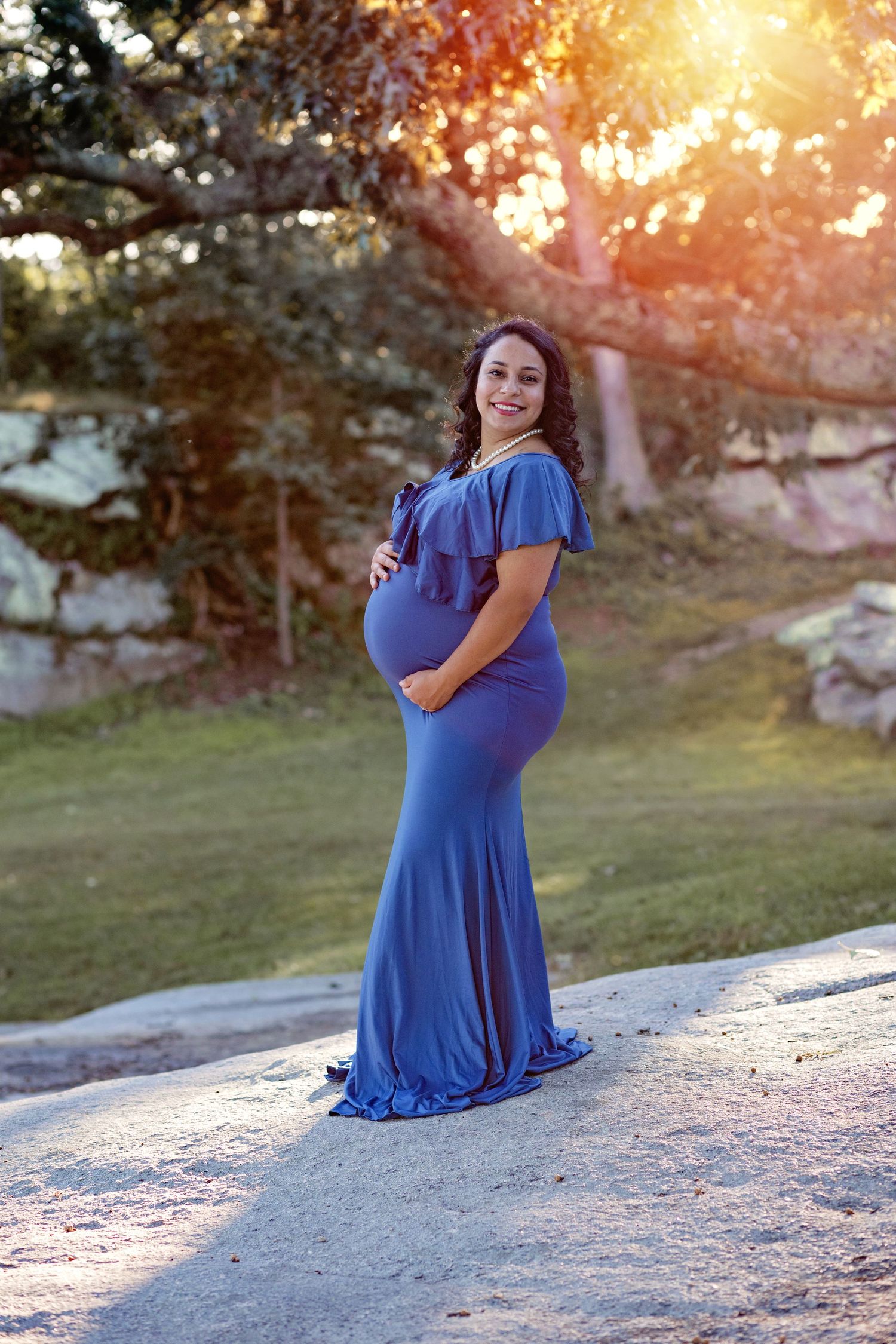A woman in a royal blue maternity dress poses on a stone ledge during golden hour sunset.
