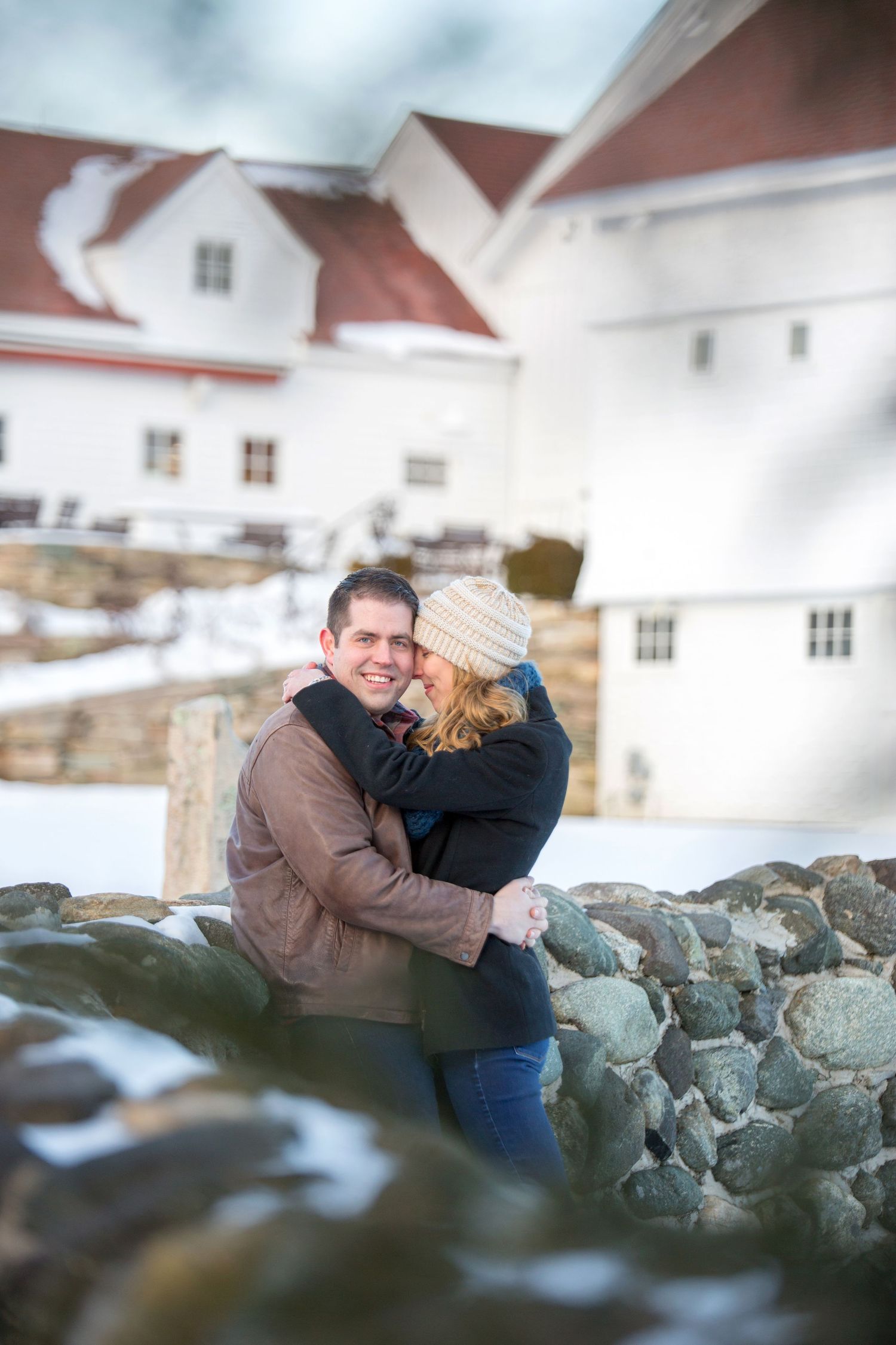 Romantic winter engagement photo series captured on rocky coastal terrain with white buildings in background.