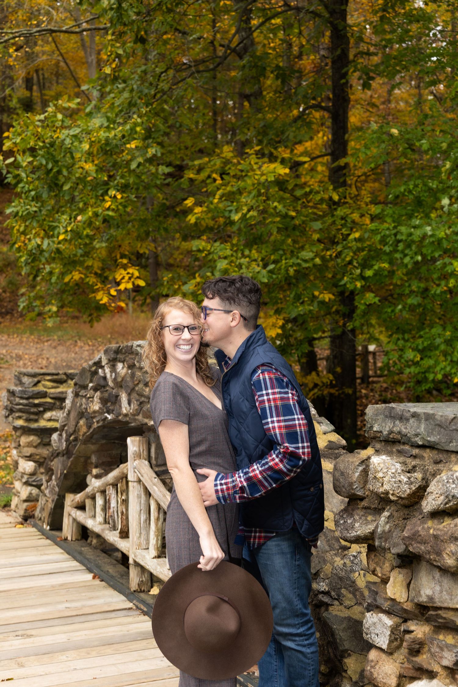 A couple shares a romantic moment on a wooden bridge surrounded by autumn foliage and stone walls.