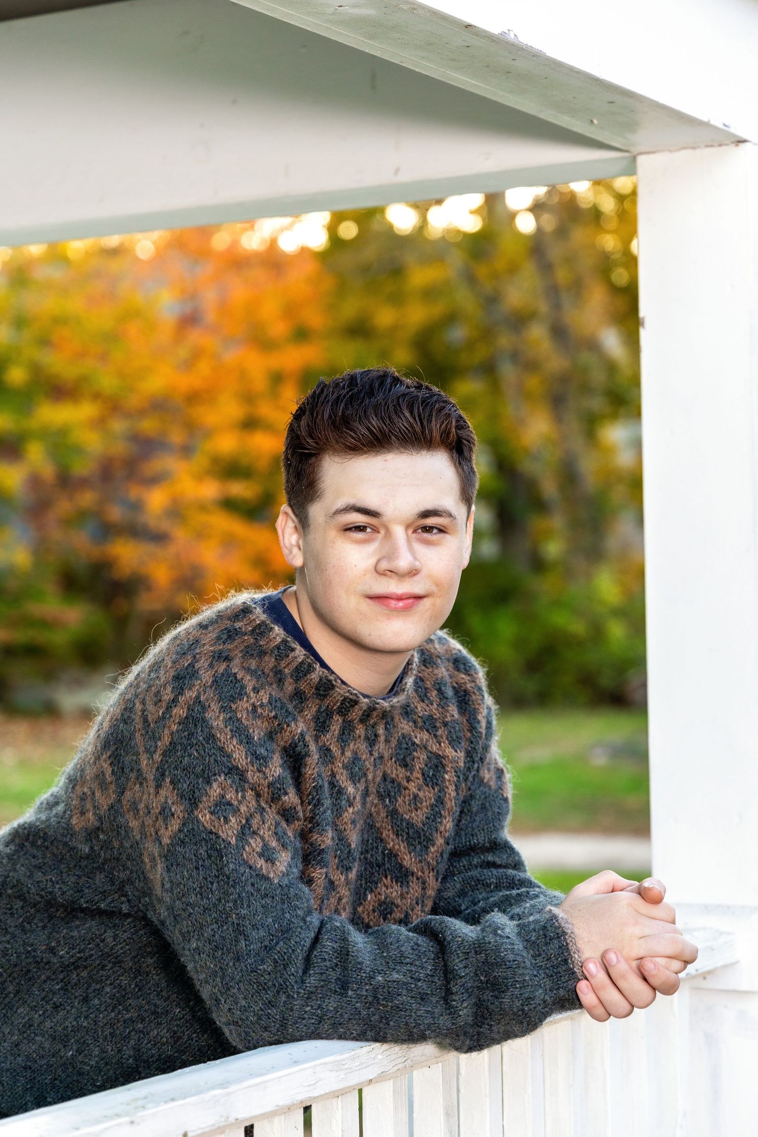 A person wearing a dark patterned sweater leans on a white railing with autumn foliage in the background.