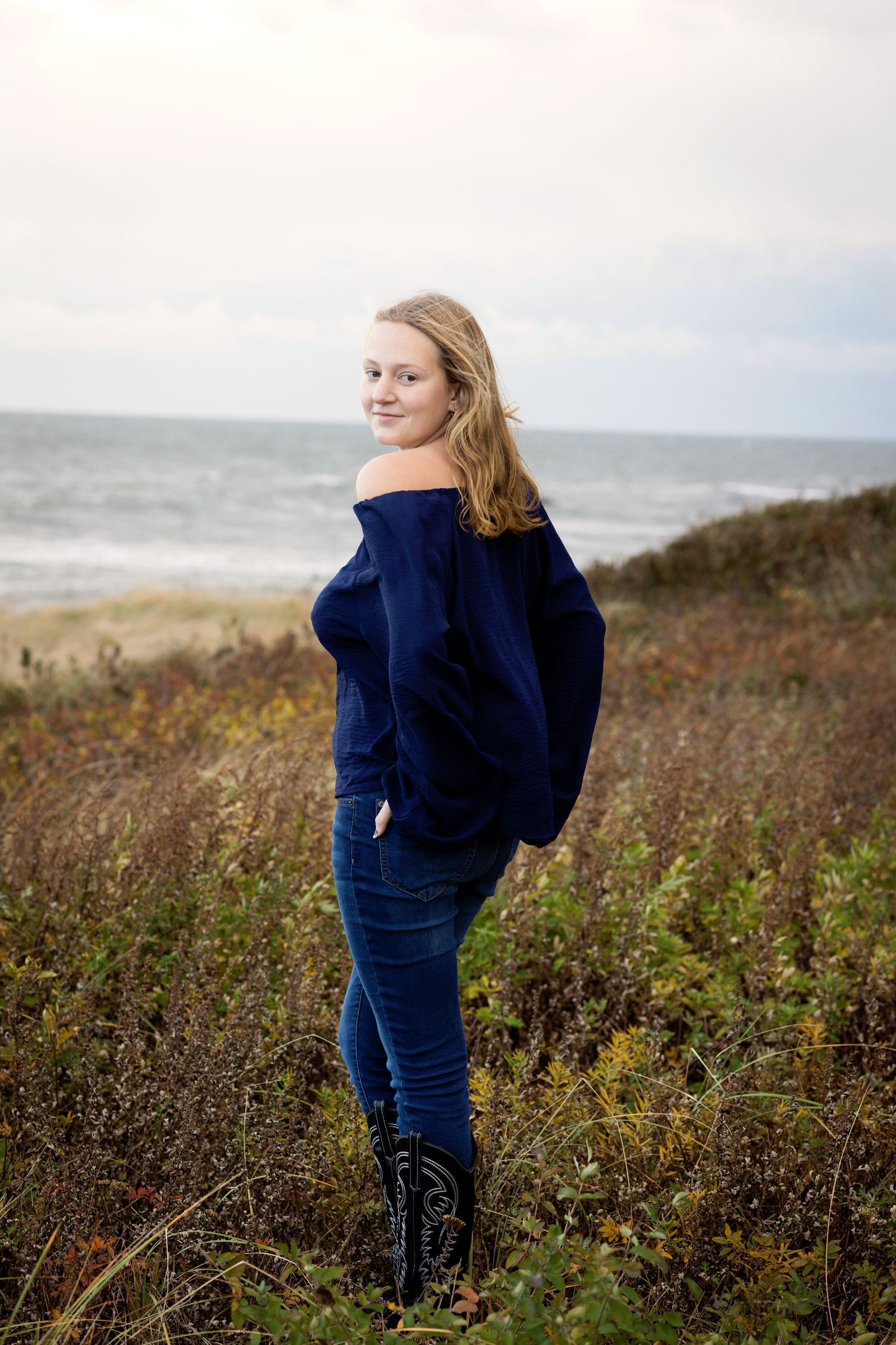 A figure in a navy sweater stands among coastal wildflowers on a foggy, windswept cliff overlooking the ocean.