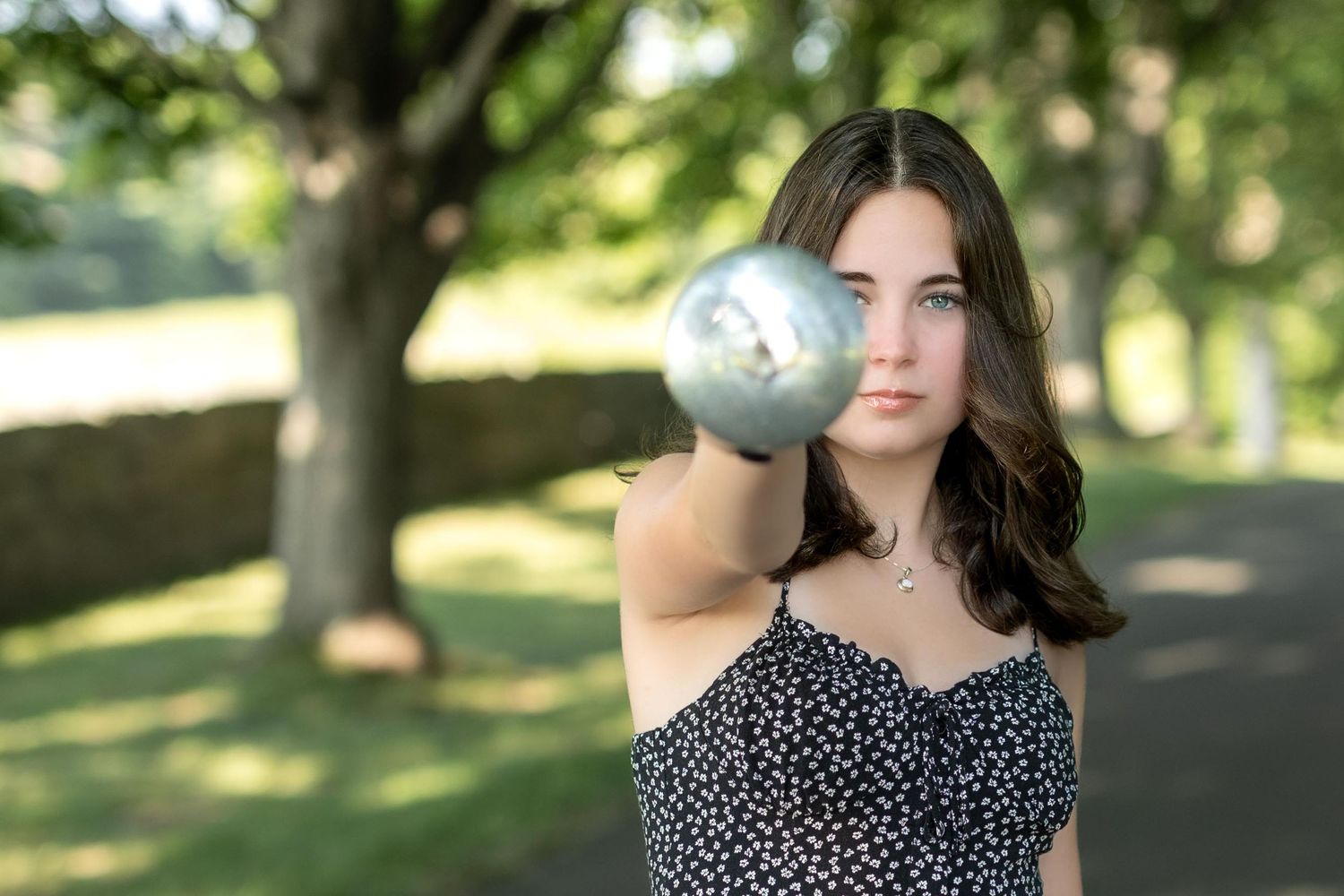 A woman in a polka dot dress poses outdoors in a park with sunlit trees creating a dreamy summer atmosphere.