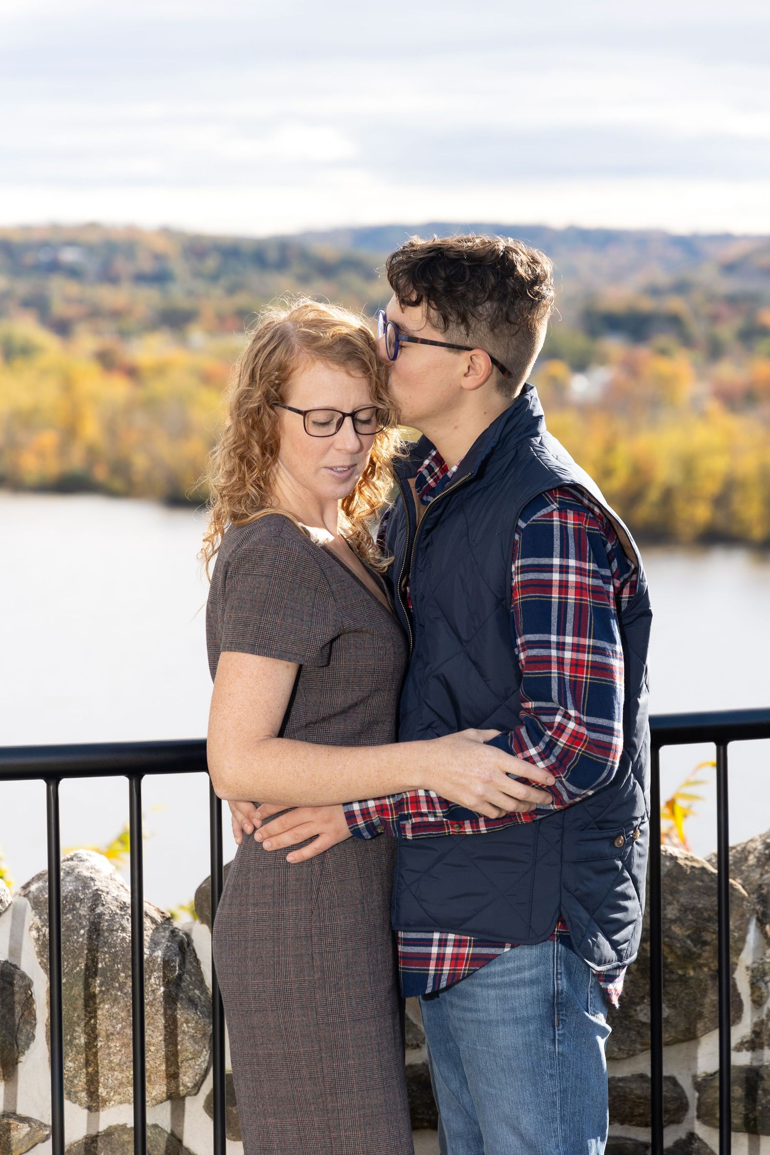 A couple embraces on a scenic overlook with autumn foliage and a lake in the background.