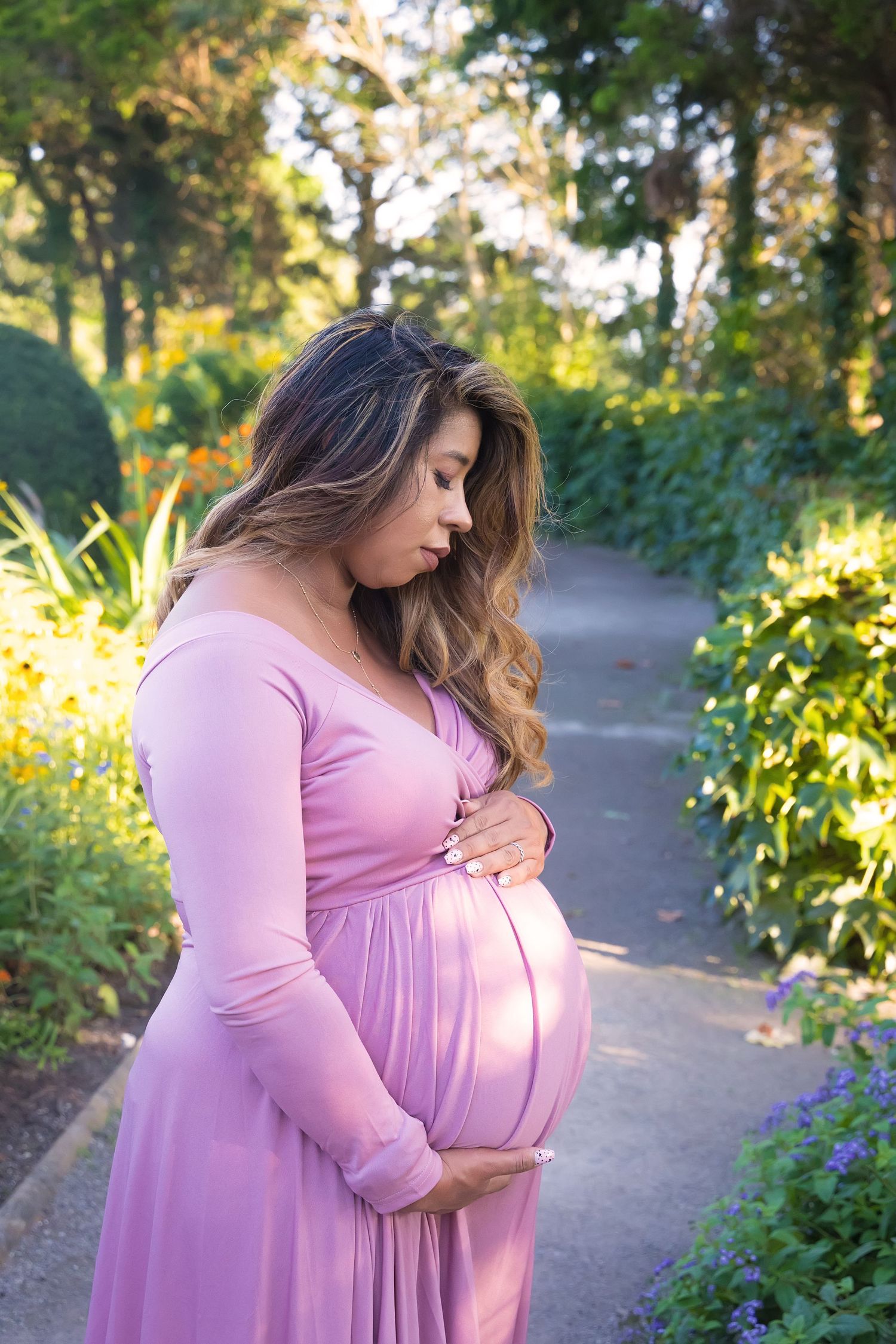 A pregnant woman in a flowing pink dress stands on a garden path surrounded by lush greenery in warm sunlight.