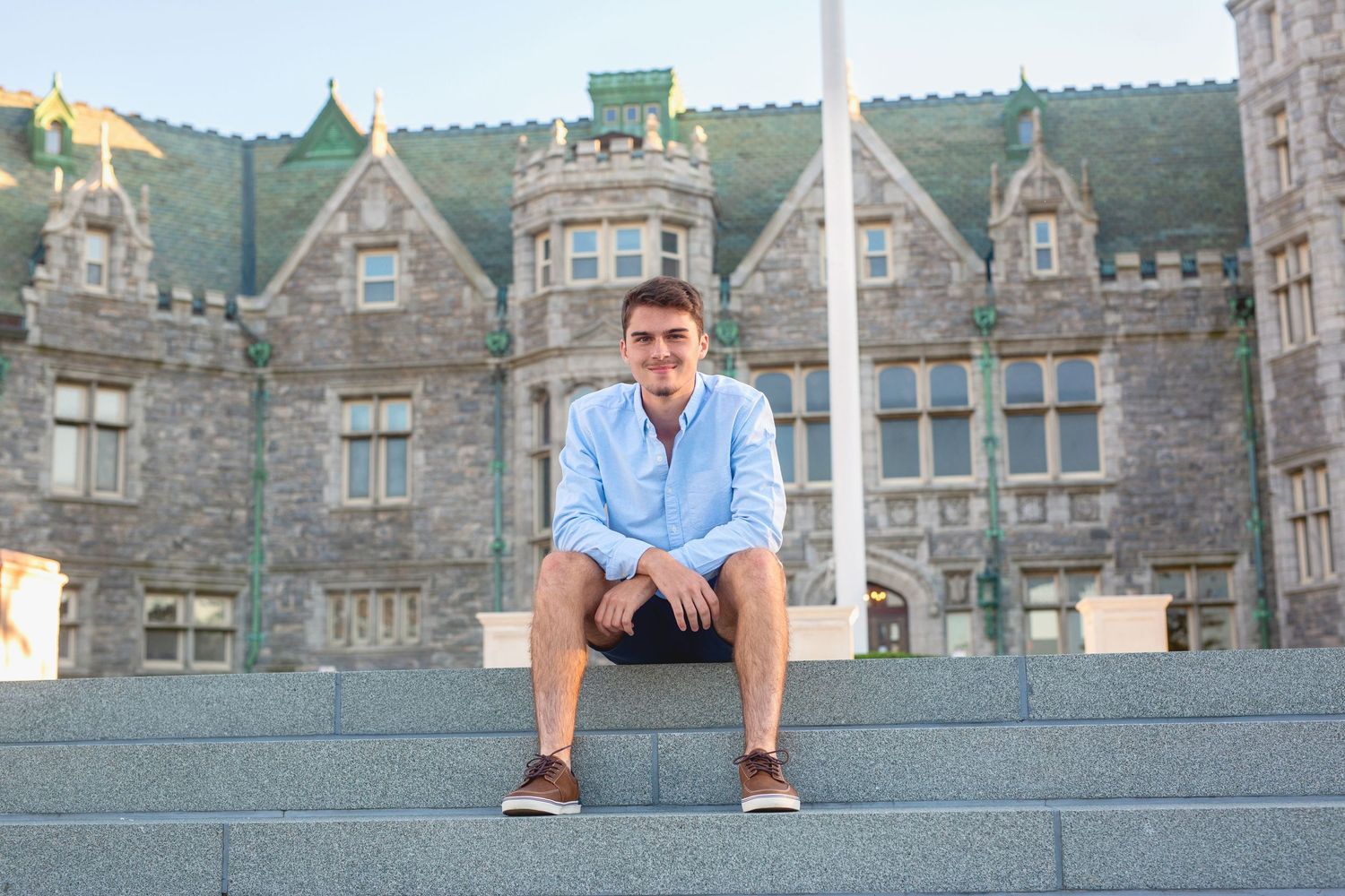 Someone sits on stone steps in front of a grand gothic stone castle or university building on a sunny day.