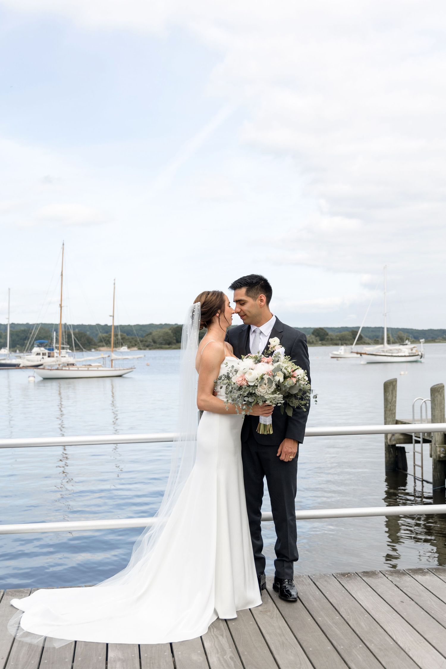 A couple shares a romantic moment on a wooden dock with sailboats and calm waters in the background.