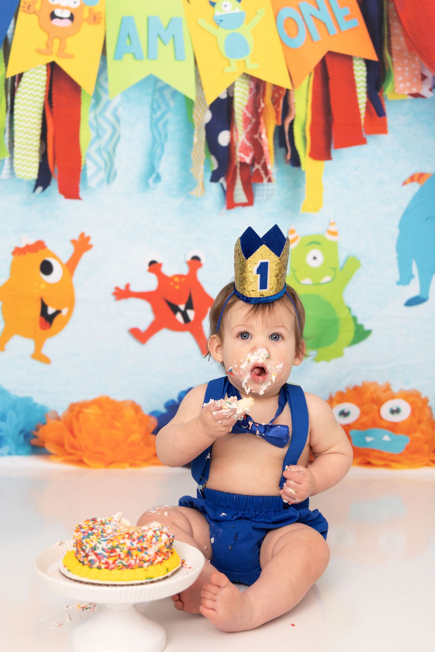 Colorful cake smash photo series with monster themed decorations and a blue outfit against festive party backdrop.
