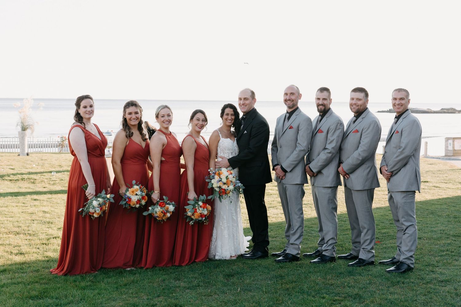 Wedding party in red dresses and gray suits posed together on oceanfront lawn.