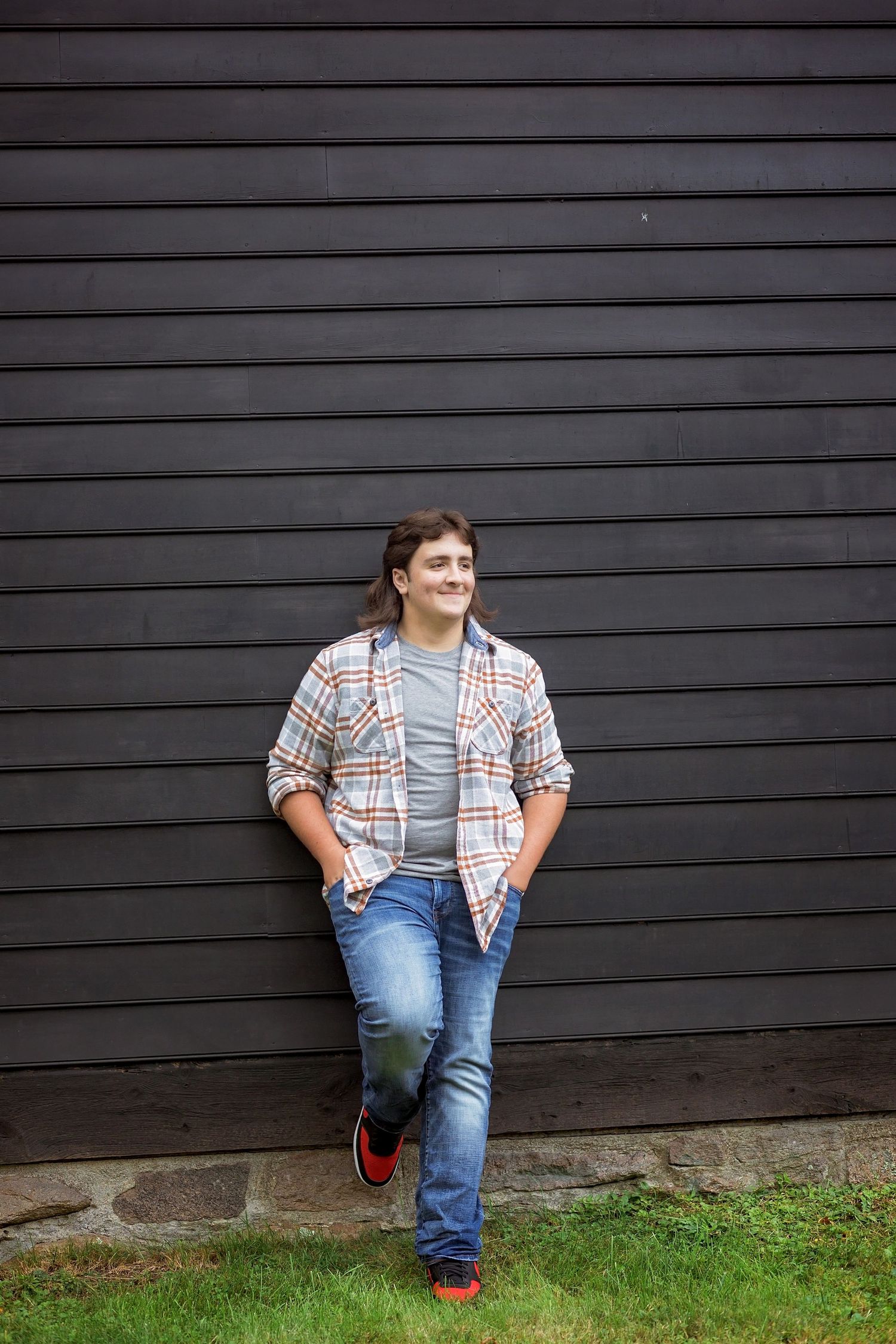 A child in plaid shirt and jeans sits casually against a dark wall with grass below.