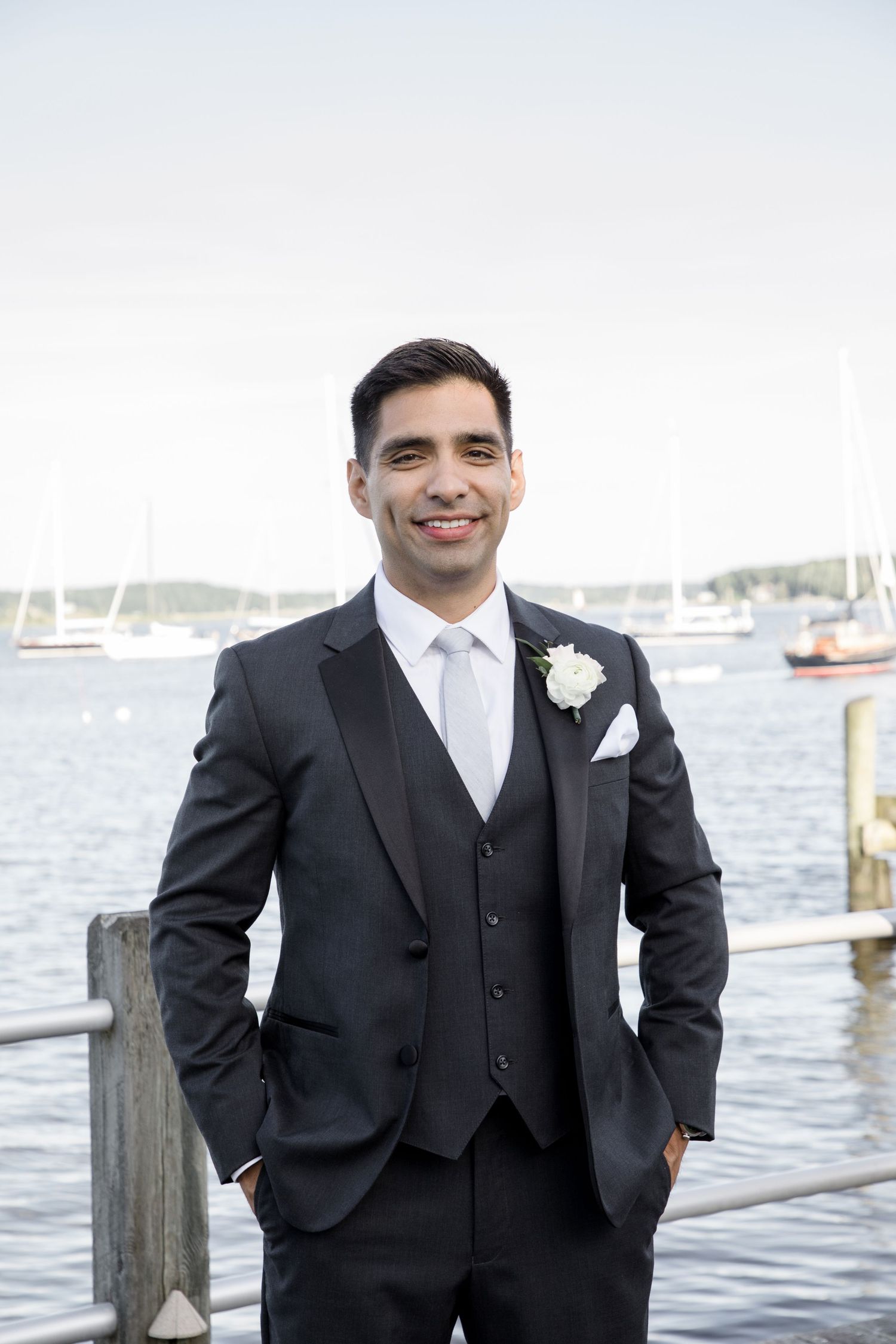 A groom in a classic black three-piece suit poses on a dock near a marina on his wedding day.