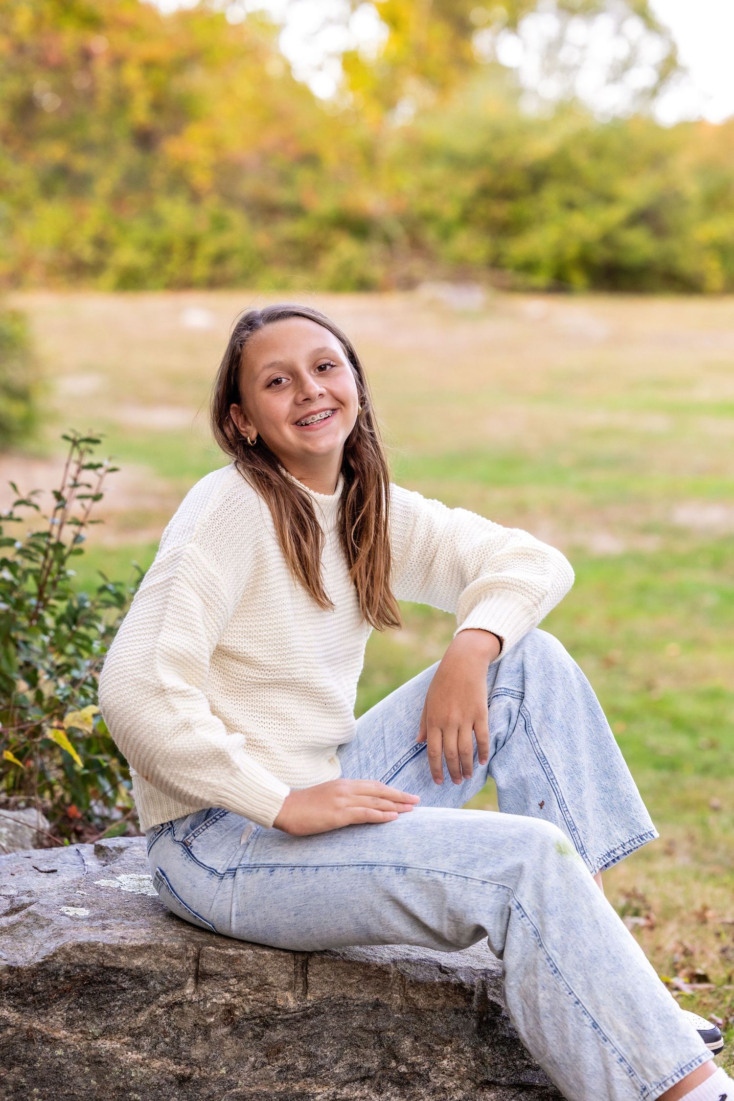 Person in white sweater and light jeans sits casually on rustic stone wall during autumn golden hour.