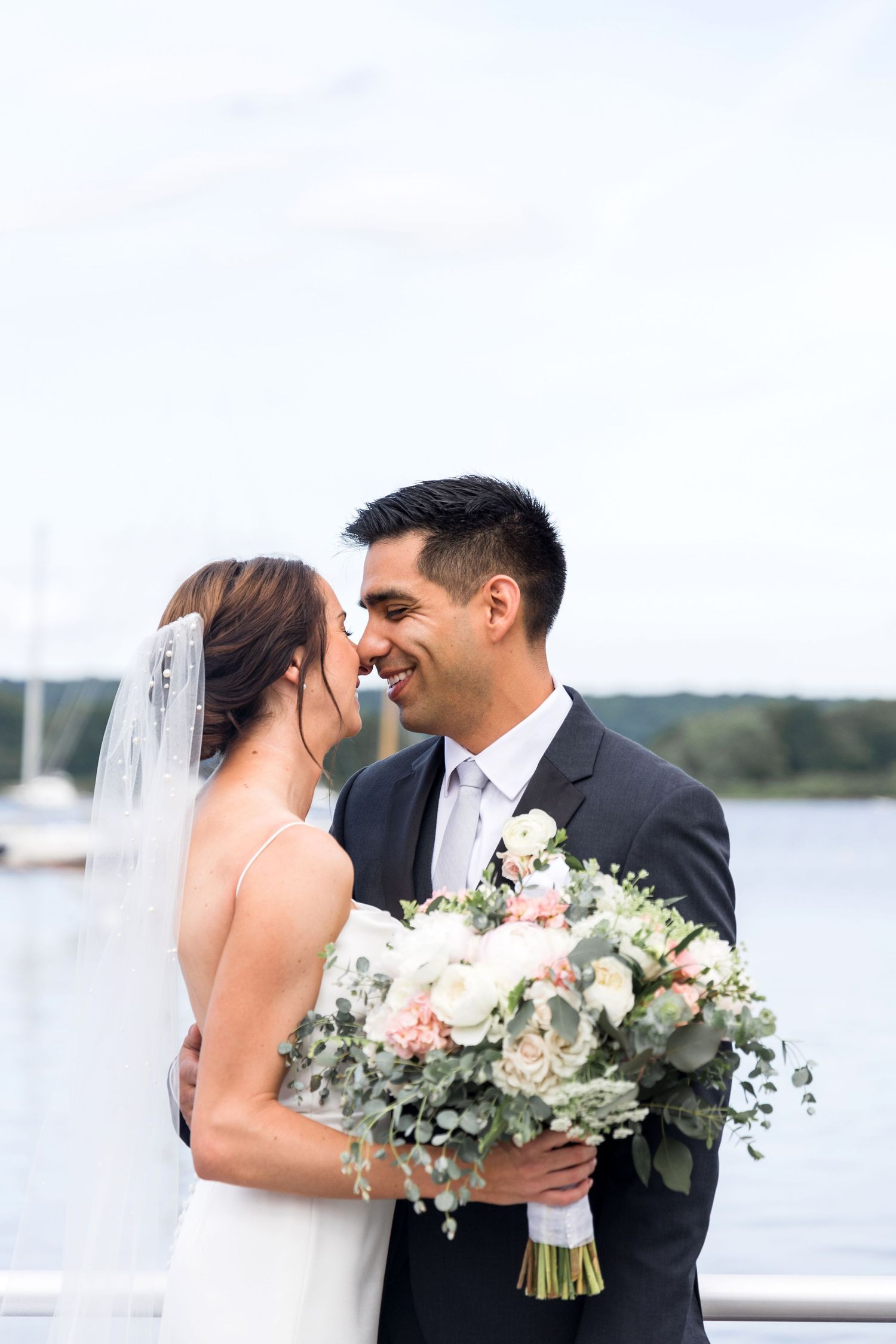 Romantic wedding couple shares intimate moments by a waterfront with a beautiful floral bouquet in soft pink and white tones.